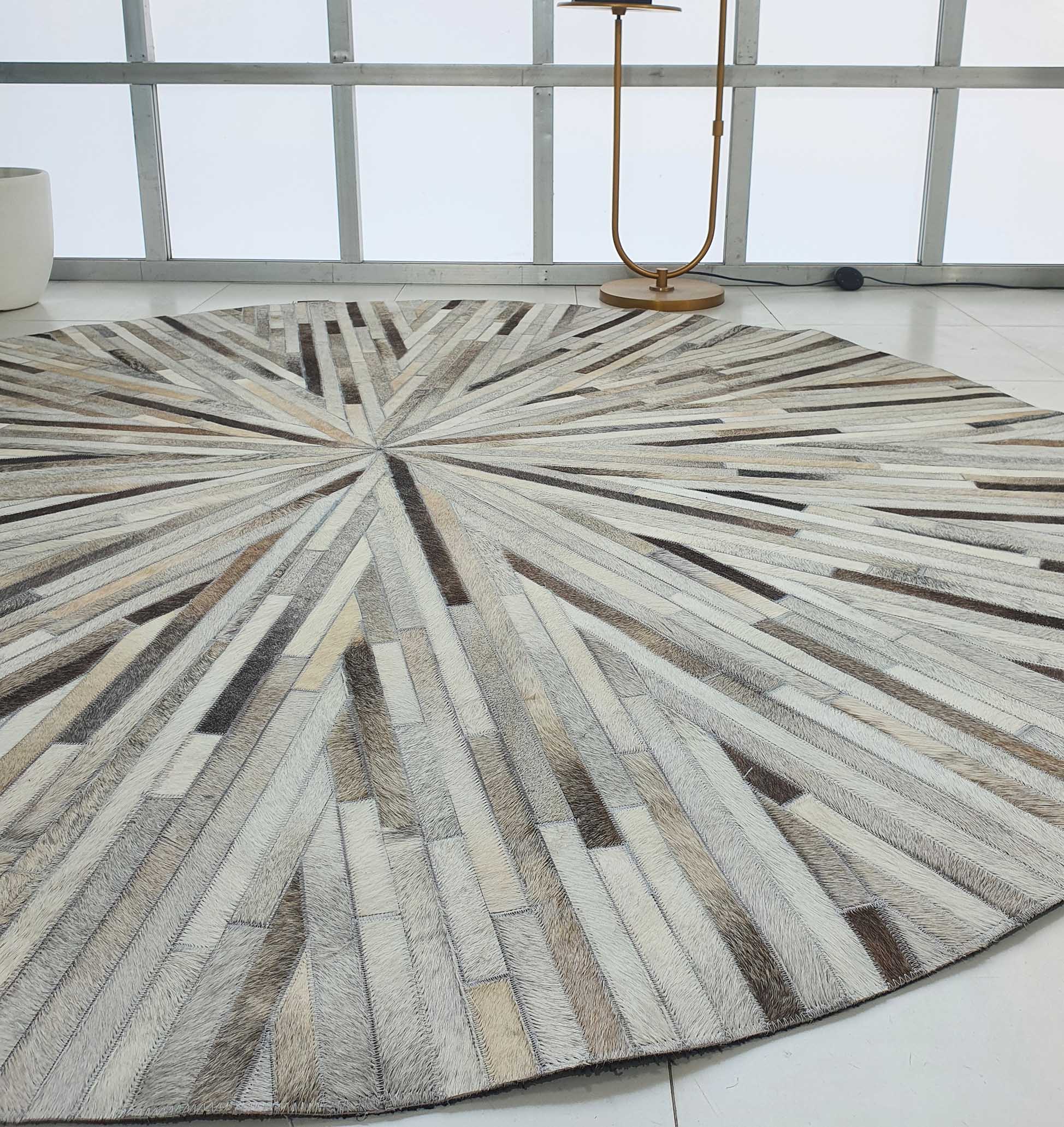 The RAMSHA HOME Home Decorative Leather Rug - LR 018 features a round starburst pattern in beige, brown, and gray segments, shown on a white tiled floor in a modern room with large windows and a gold floor lamp.