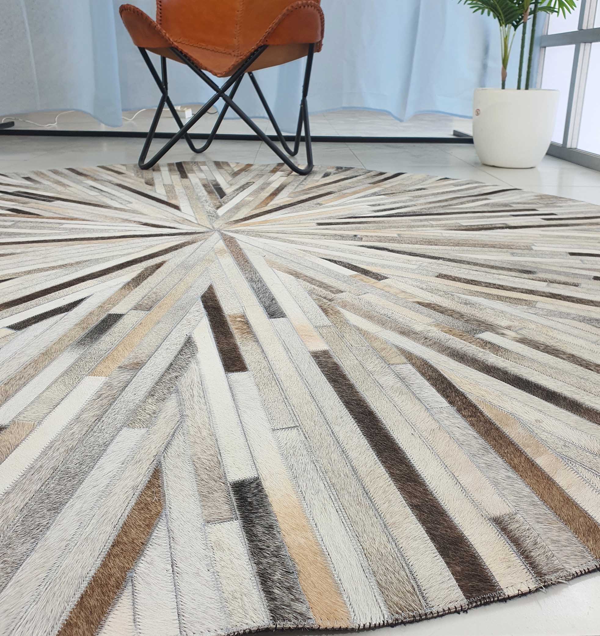 The RAMSHA HOME Home Decorative Leather Rug - LR 018 features a sunburst pattern in neutral shades, covering the floor. In the background, a brown leather chair sits near a white potted plant and a window with sheer curtains.