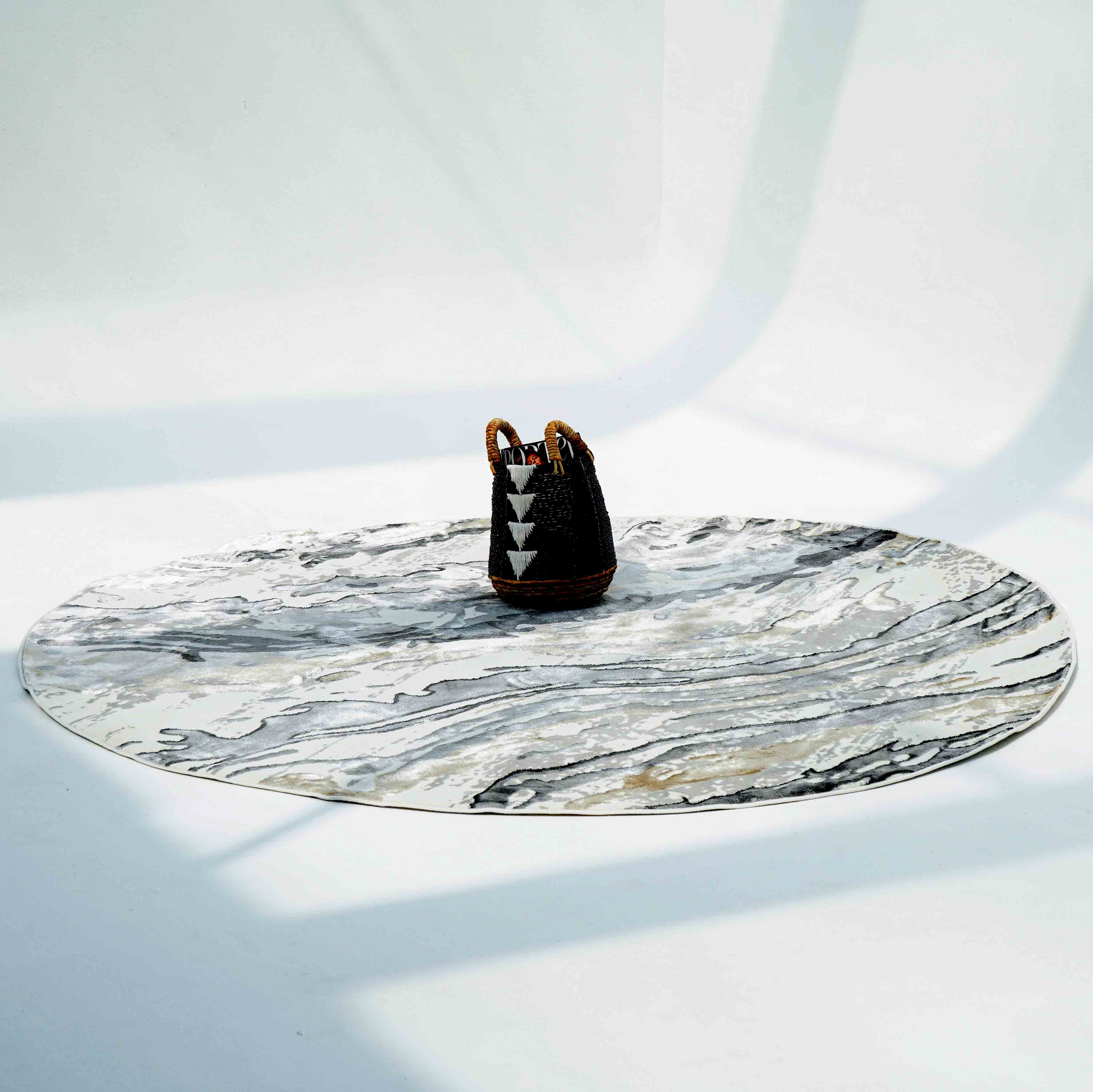 The Shrink Cream ND53K Polyester Decorative Floor Rug by RAMSHA HOME, featuring a large round marble pattern, is softly illuminated with a small black and brown woven bag with white triangles placed on top.