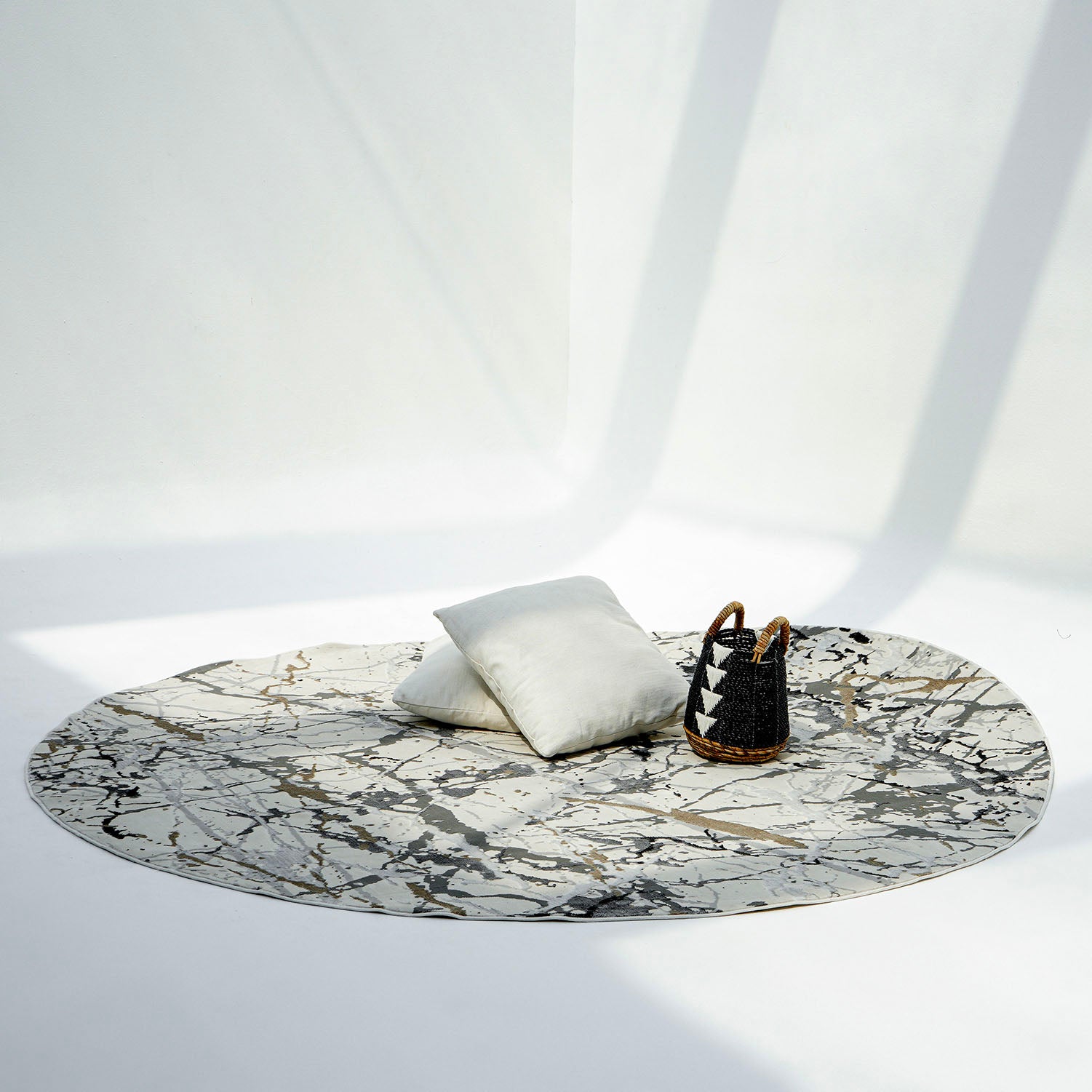 A minimalist scene with a cream-colored RAMSHA HOME Machine Made Turkey Carpet MZ29C, two light pillows, and a black-and-brown basket on a white floor bathed in soft natural light and shadows.