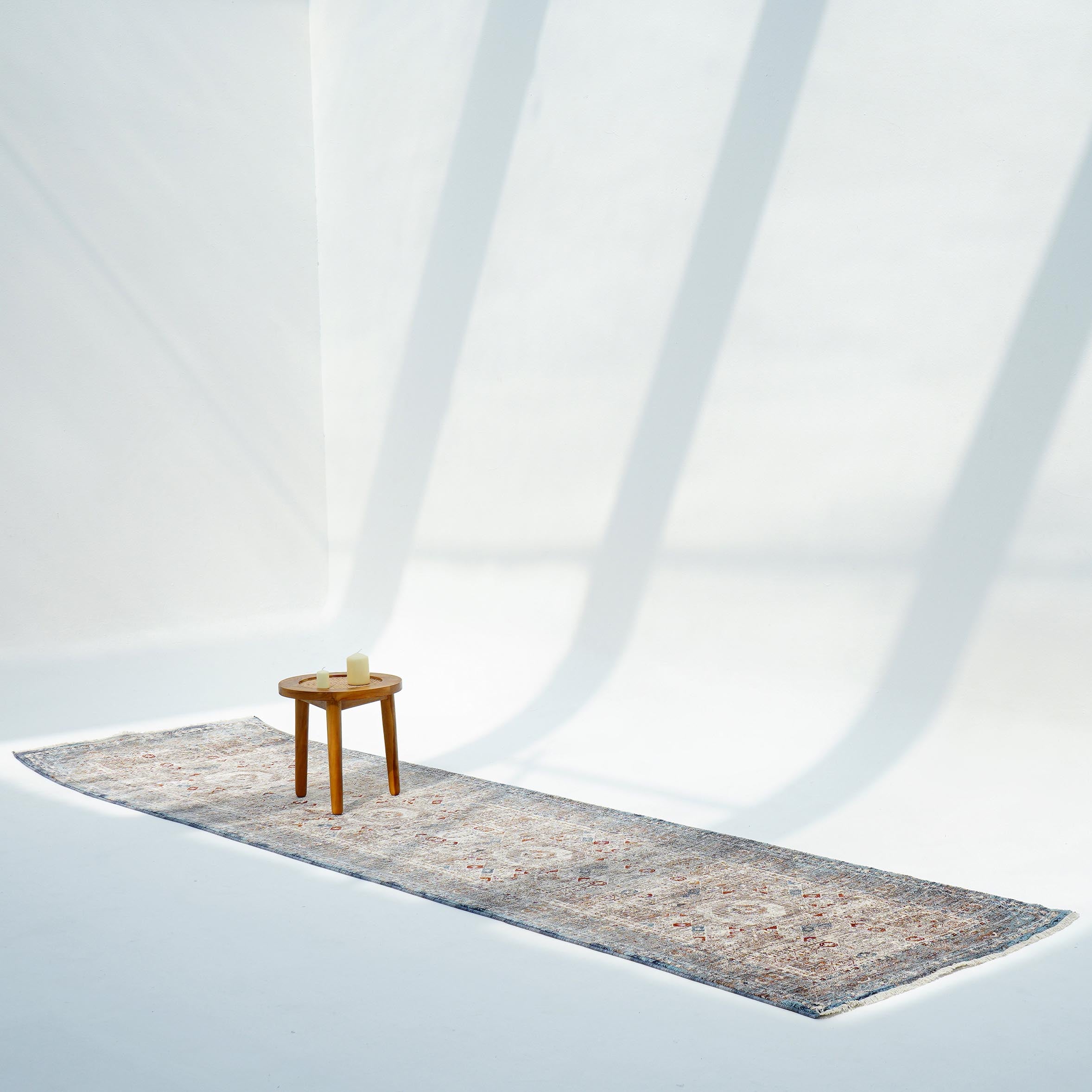 A small wooden stool with a candle sits on the RAMSHA HOME Polyester K860A Area Rug in a bright, minimalist room, where long shadows fall across white walls and the floor.