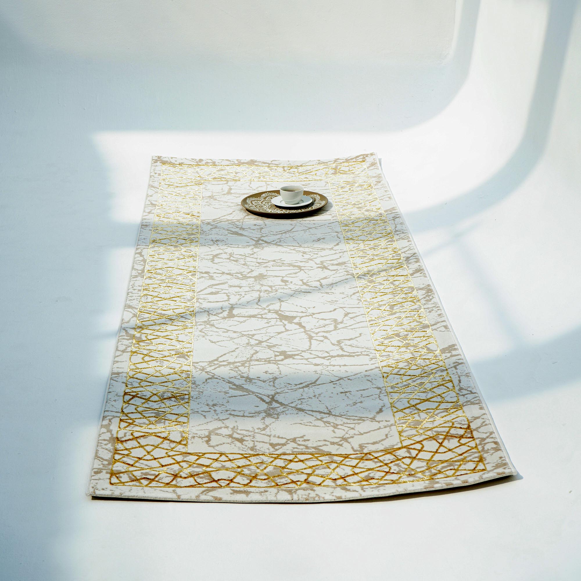 The RAMSHA HOME Cream, Shrink Cream Colored Geometry Pattern Turkey Rug MZ67C lies on a white floor, with a cup and saucer on a decorative plate near the center, as natural light casts soft shadows across the scene.