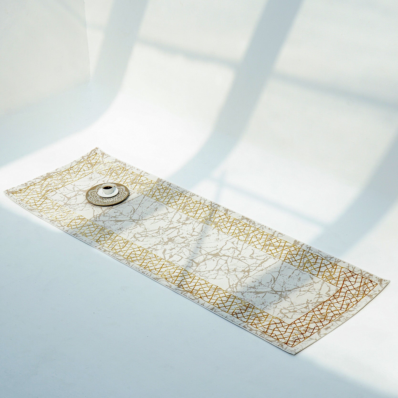 The RAMSHA HOME Cream, Shrink Cream Colored Geometry Pattern Turkey Rug MZ67C showcases a geometric design and marble-like center in cream and ivory tones on a white floor, with sunlight casting window shadows across a teacup and saucer in the corner.