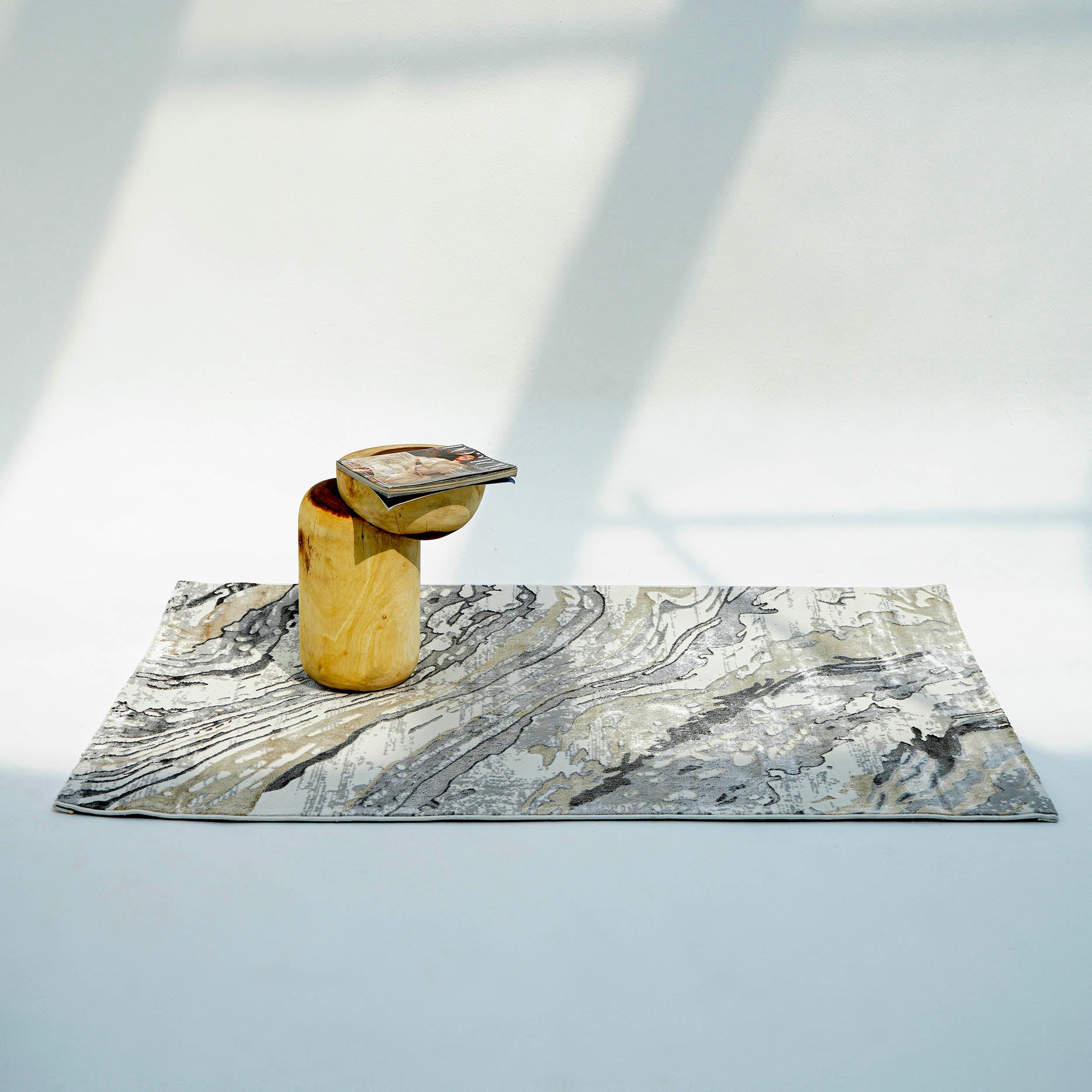 The RAMSHA HOME Shrink Cream ND53K Polyester Decorative Floor Rug with a marble pattern lies on a light floor, accented by a small cylindrical wooden side table holding a magazine as sunlight casts soft shadows across the scene.