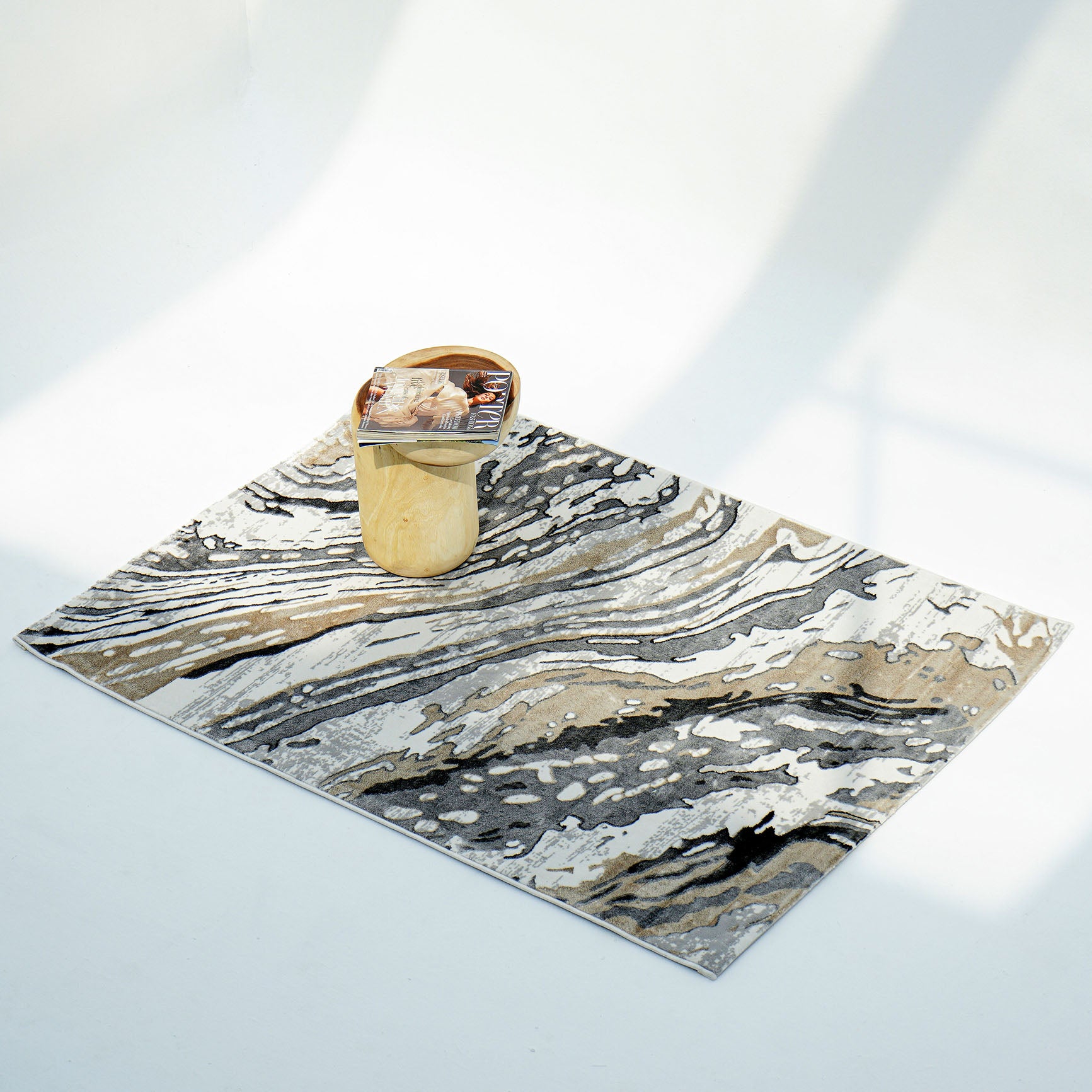 The RAMSHA HOME Shrink Cream ND53K Polyester Decorative Floor Rug, featuring modern gray beige marble-like abstract patterns, rests on a white floor. A light wood side table with magazines sits atop the rug amid soft shadows.