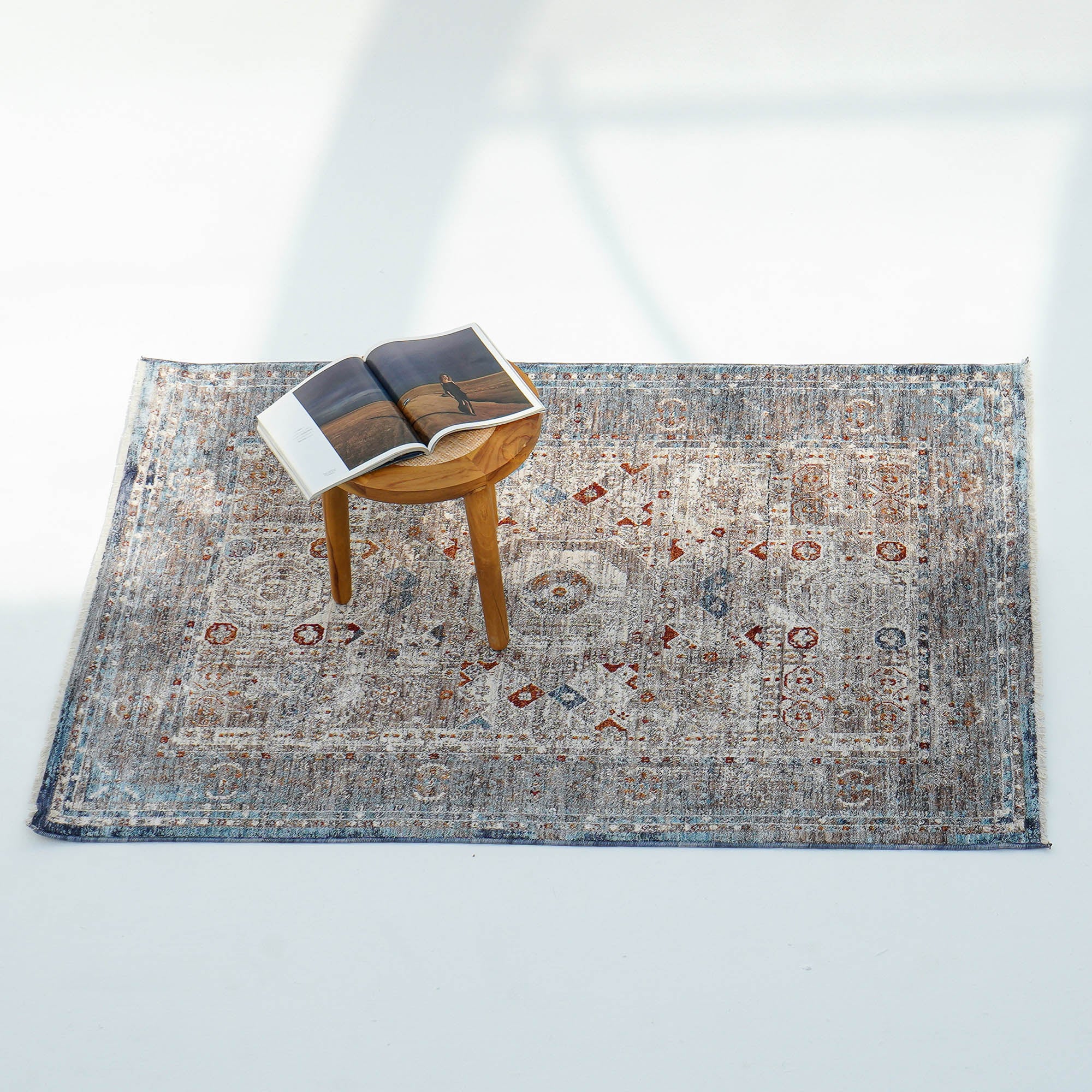 The RAMSHA HOME Polyester K860A Area Rug, featuring blue and red accents, lies on a white floor. A wooden stool with an open photo book sits on the rug as sunlight creates soft shadows in this tranquil living space.
