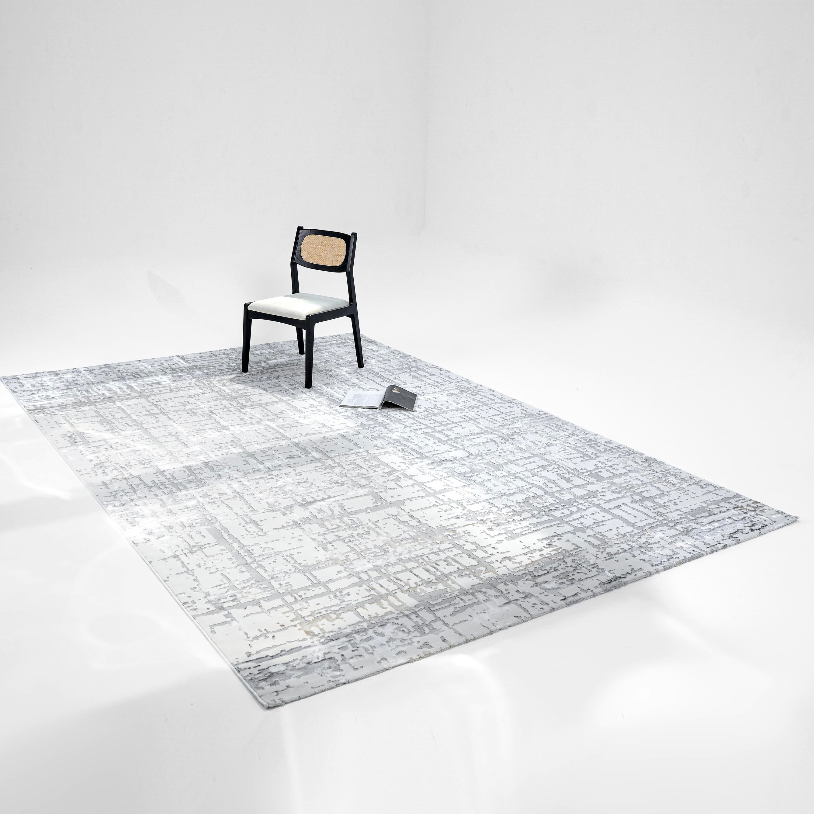 A black chair with a white seat and woven backrest sits on the RAMSHA HOME Cream Colored Geometry Turkish Carpet MZ24Z in a minimal white room. A closed book rests nearby on the patterned surface of the modern rug.