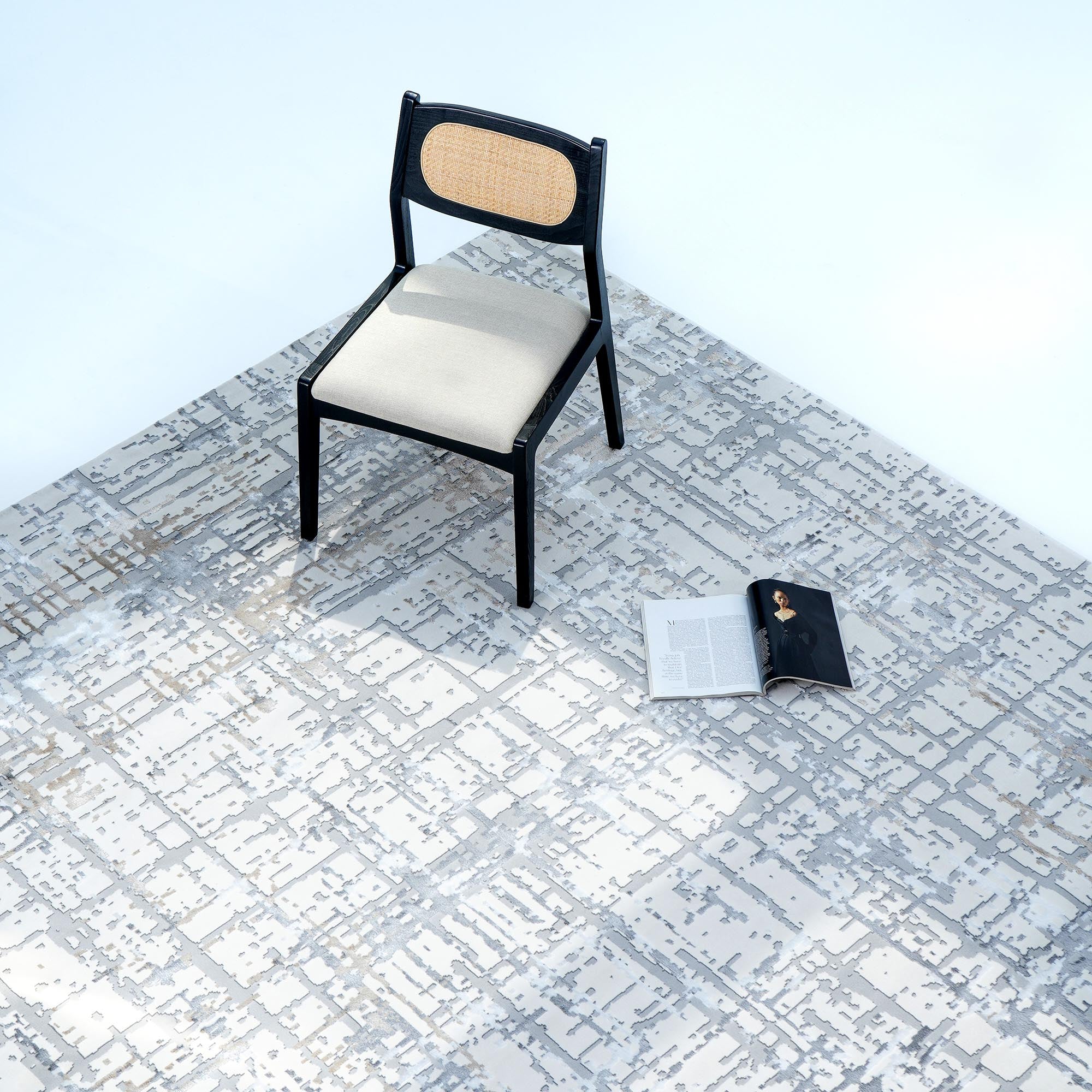 A black chair with a light cushion and woven backrest sits atop the RAMSHA HOME Cream Colored Geometry Turkish Carpet MZ24Z, on a geometric floor. An open magazine rests nearby, enhancing the minimal, well-lit scene.