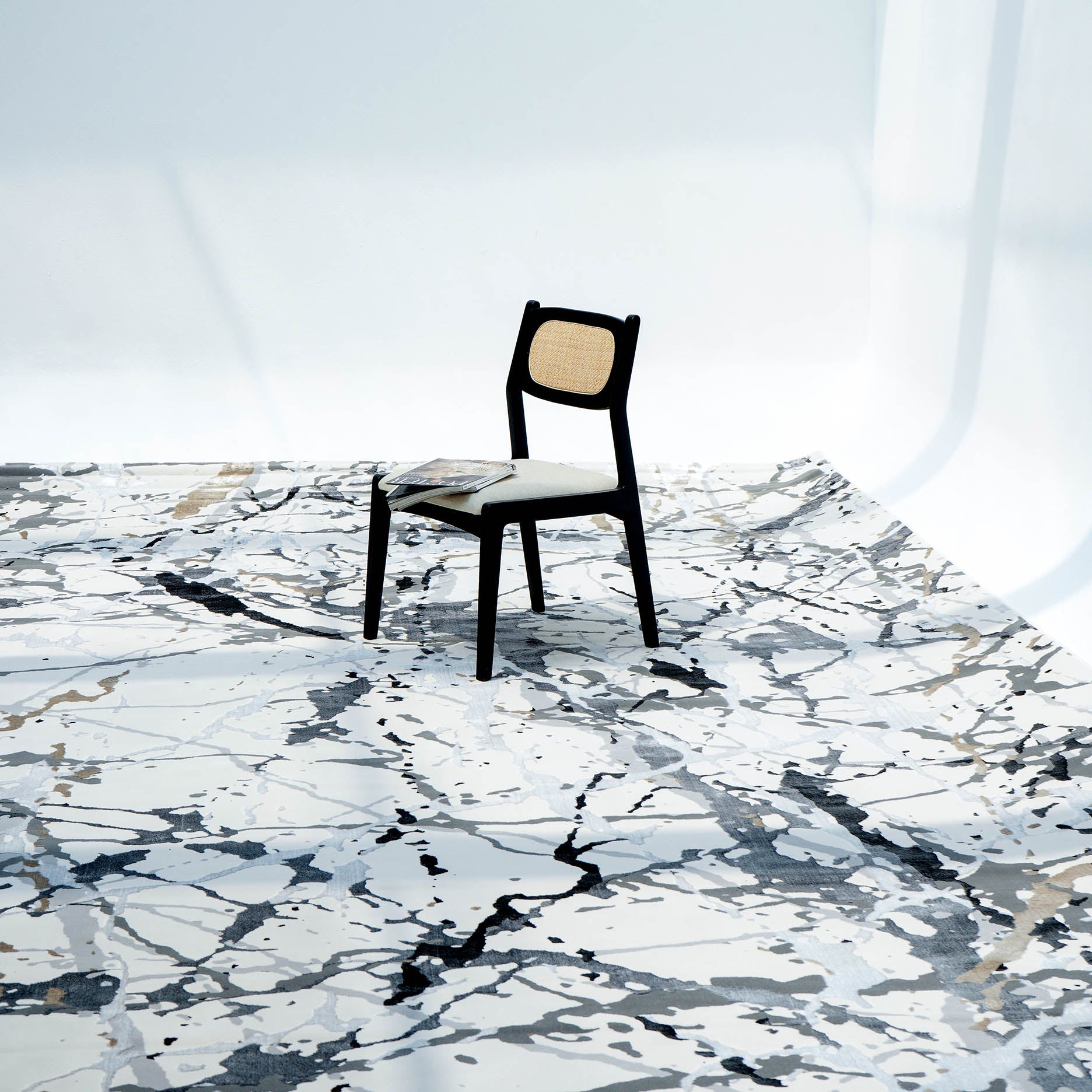 A single black chair with a light seat and backrest sits on the RAMSHA HOME Machine Made Turkey Carpet Cream Colored MZ29C, featuring bold abstract patterns in white, gray, and beige against a white minimalist background.