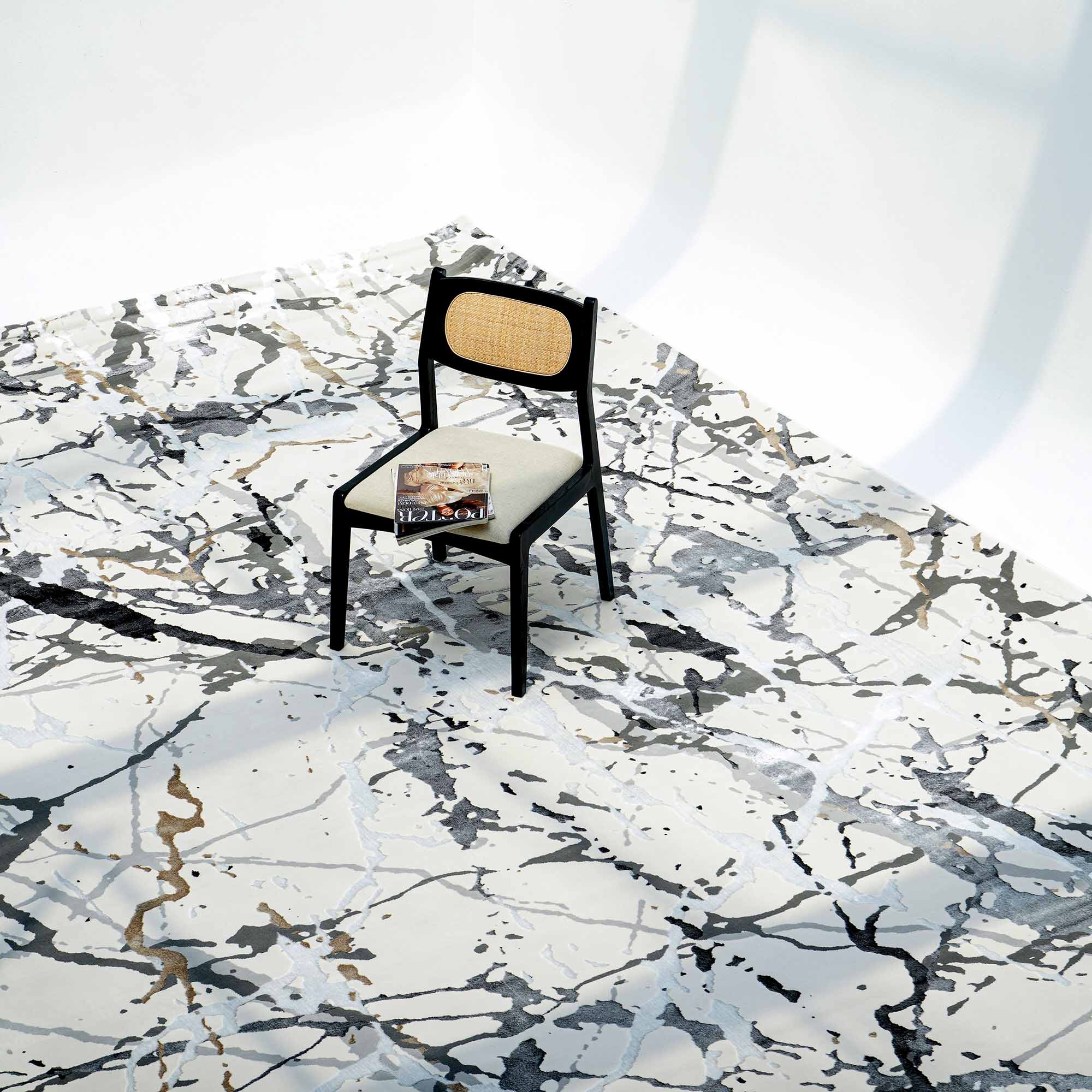 A black chair with a cane seat sits on a RAMSHA HOME Machine Made Turkey Carpet in cream (MZ29C) over a white marble-look floor. A magazine rests on the chair, all set in a minimal and well-lit space.