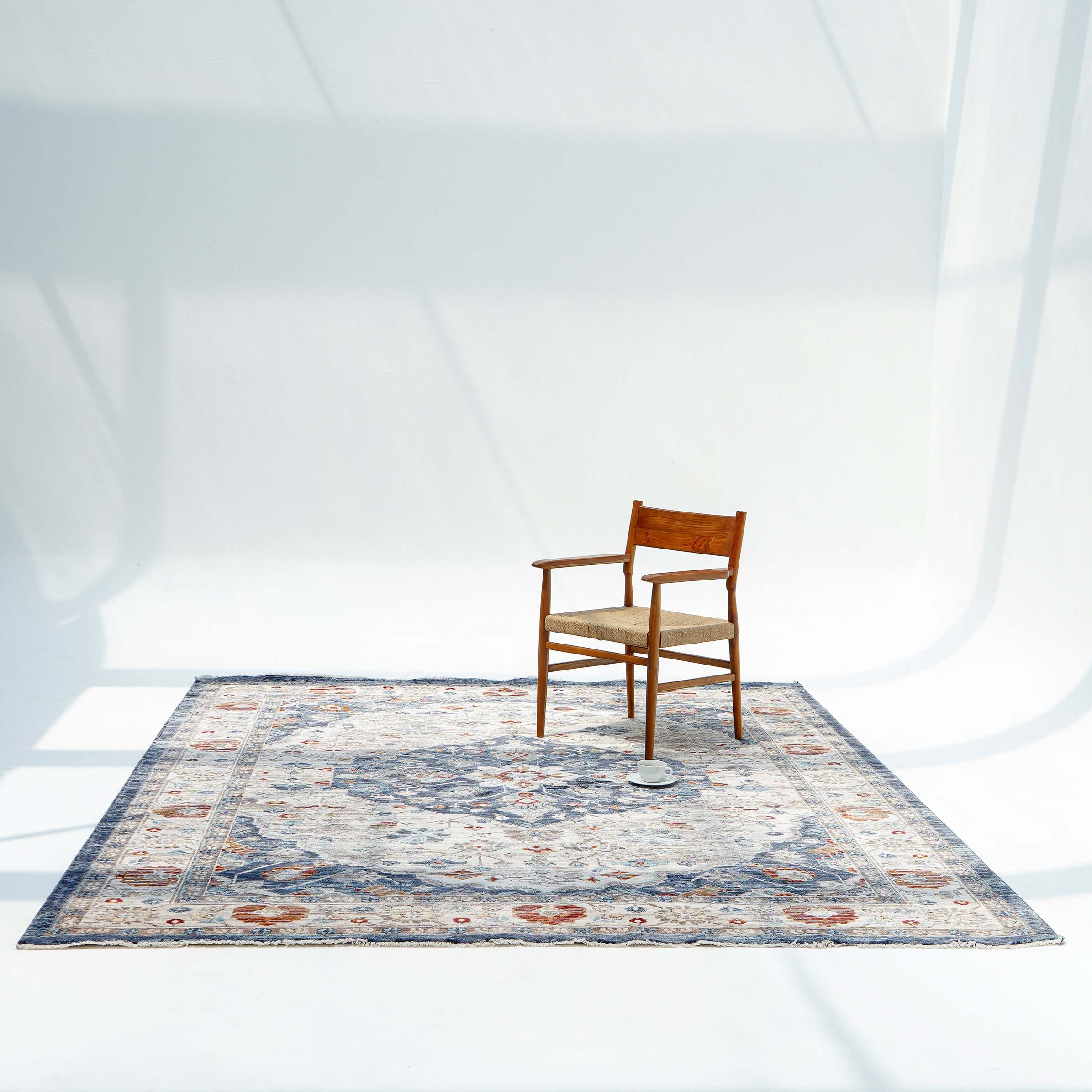 A wooden chair sits in a bright, minimal room with white walls and shadows, resting atop the RAMSHA HOME Indoor Floor Area Rug K856B 100% Polyester Silk. A small round object lies on the patterned rug near the chair.