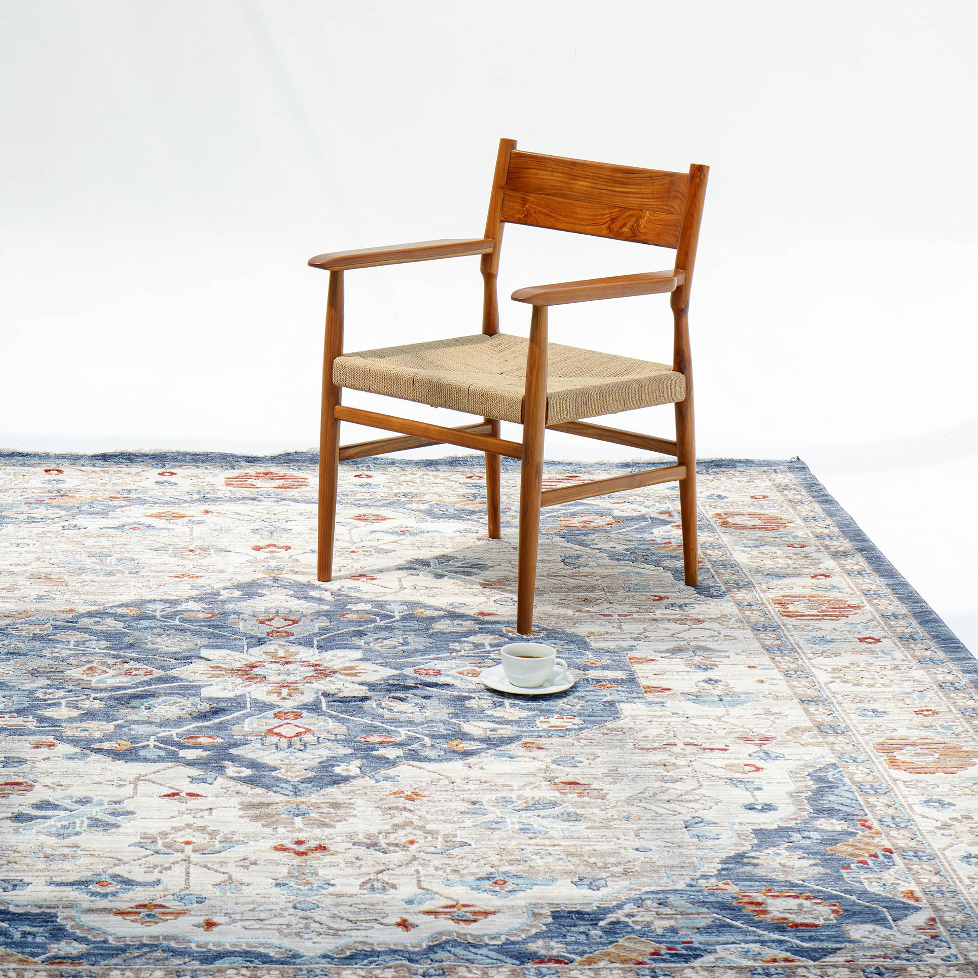 A wooden chair with a woven seat sits on the RAMSHA HOME Indoor Floor Area Rug K856B in blue and beige polyester silk, with a white cup and saucer on the floor nearby against a plain white background.