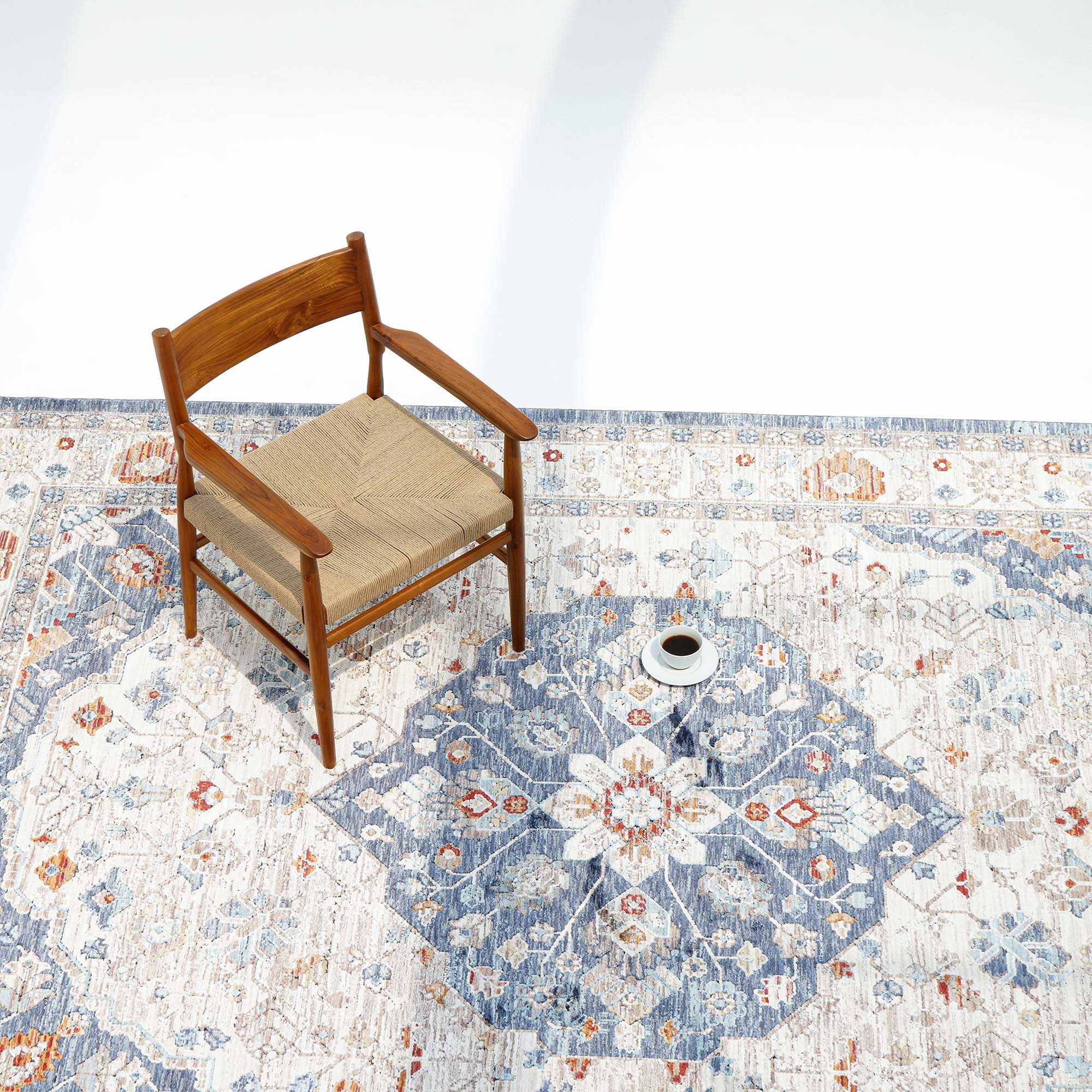 A wooden chair with a woven seat sits on the RAMSHA HOME Indoor Floor Area Rug K856B (100% Polyester Silk), featuring blue, orange, and cream medallion designs. A white cup rests nearby, all in a bright, minimalist setting.