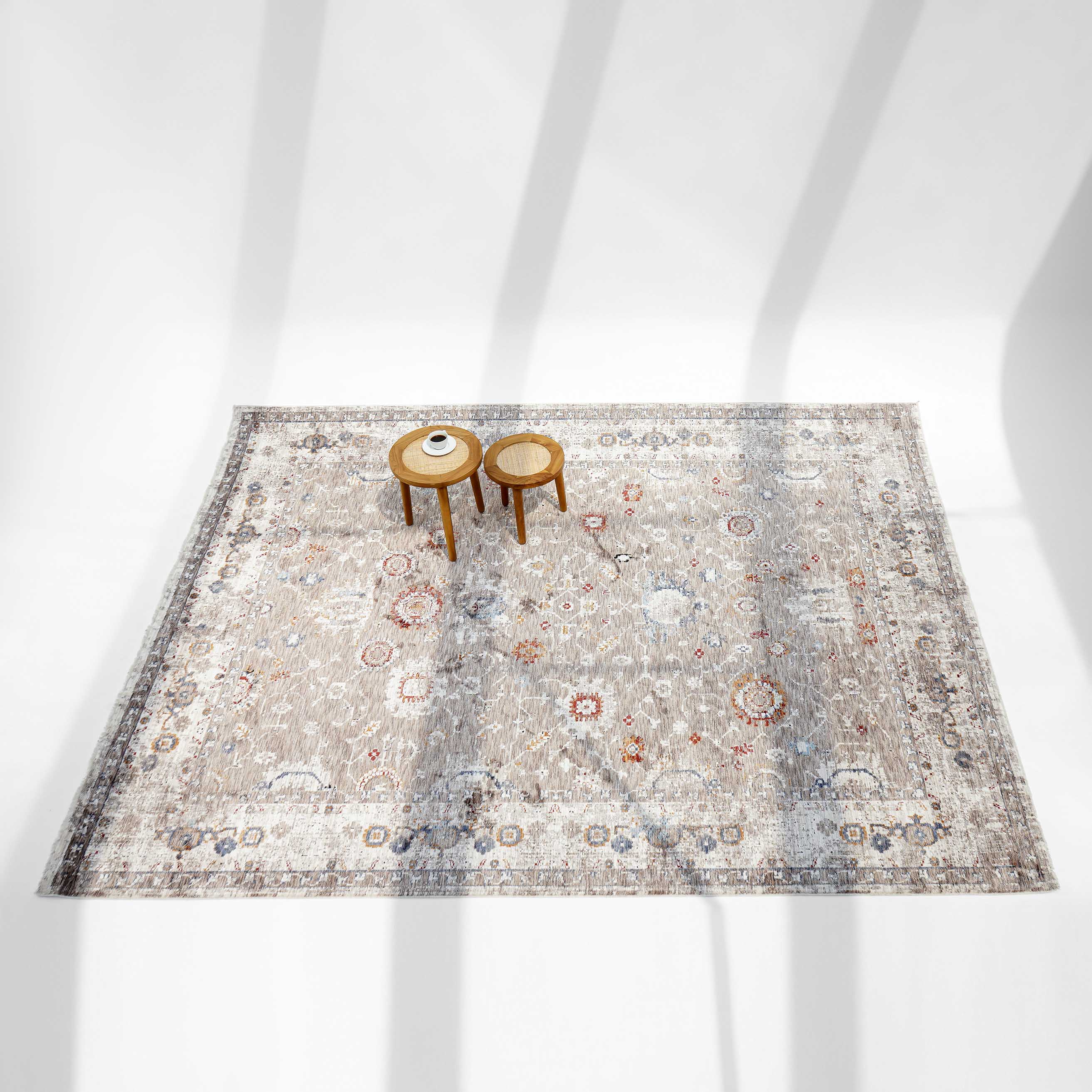 The RAMSHA HOME Beige Cream Decorative Indoor Rug K747E with floral patterns in beige, gray, and orange is shown with two round wooden side tables—one holding a cup and plate—while shadows fall across the scene.