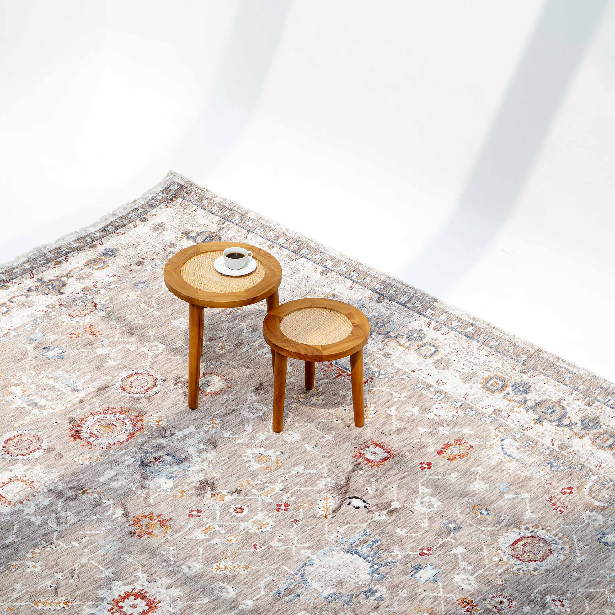 Two round wooden side tables of varying heights sit on a RAMSHA HOME Beige Cream Decorative Indoor Rug K747E. The taller table displays a white cup and saucer, while the smaller one is empty. The brightly lit scene casts minimal shadows.