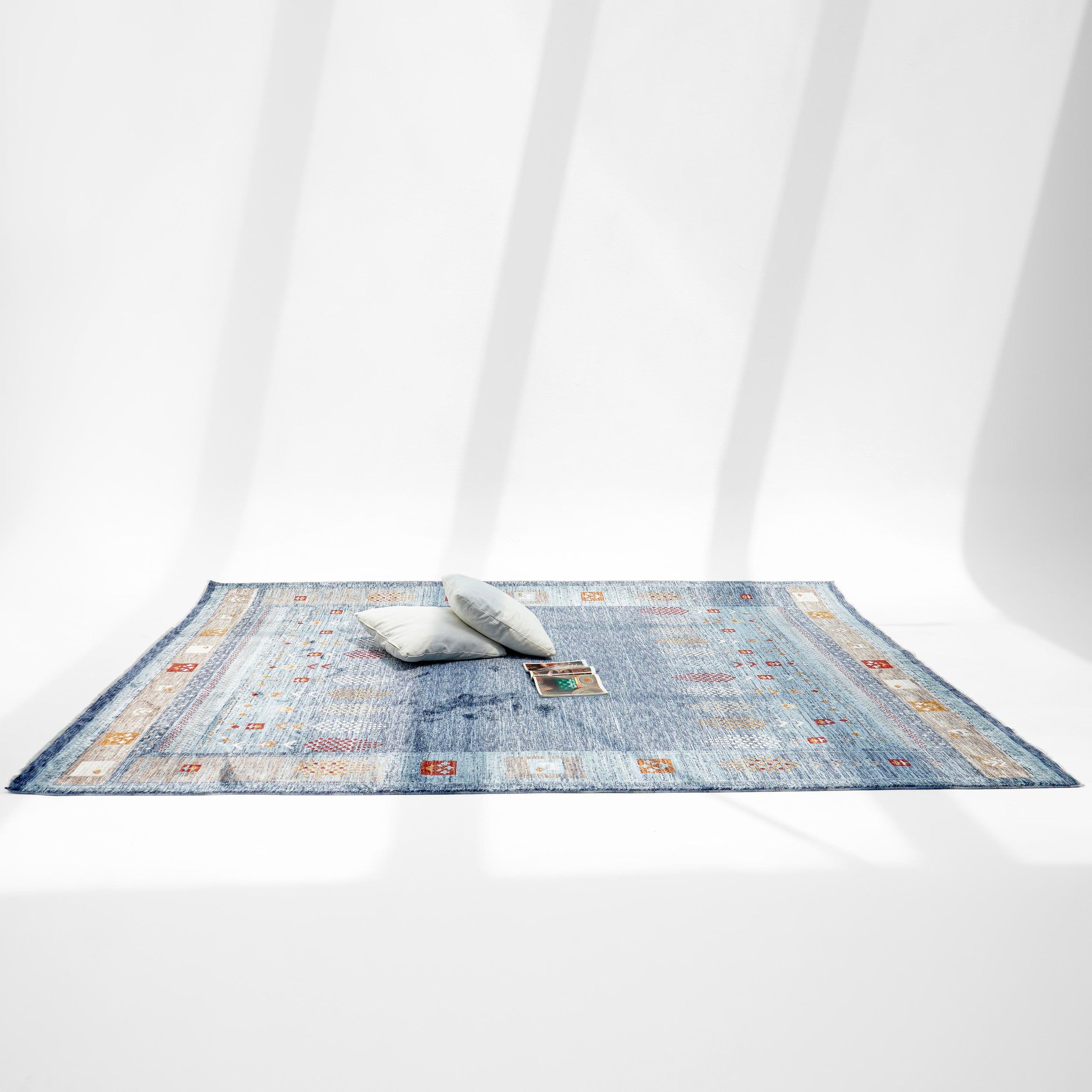 The Luxurious Denim Machine Made Turkey Rug - K742B by RAMSHA HOME, in a modern geometric blue design, is styled with two light pillows and an open book on a white floor beneath soft shadows.
