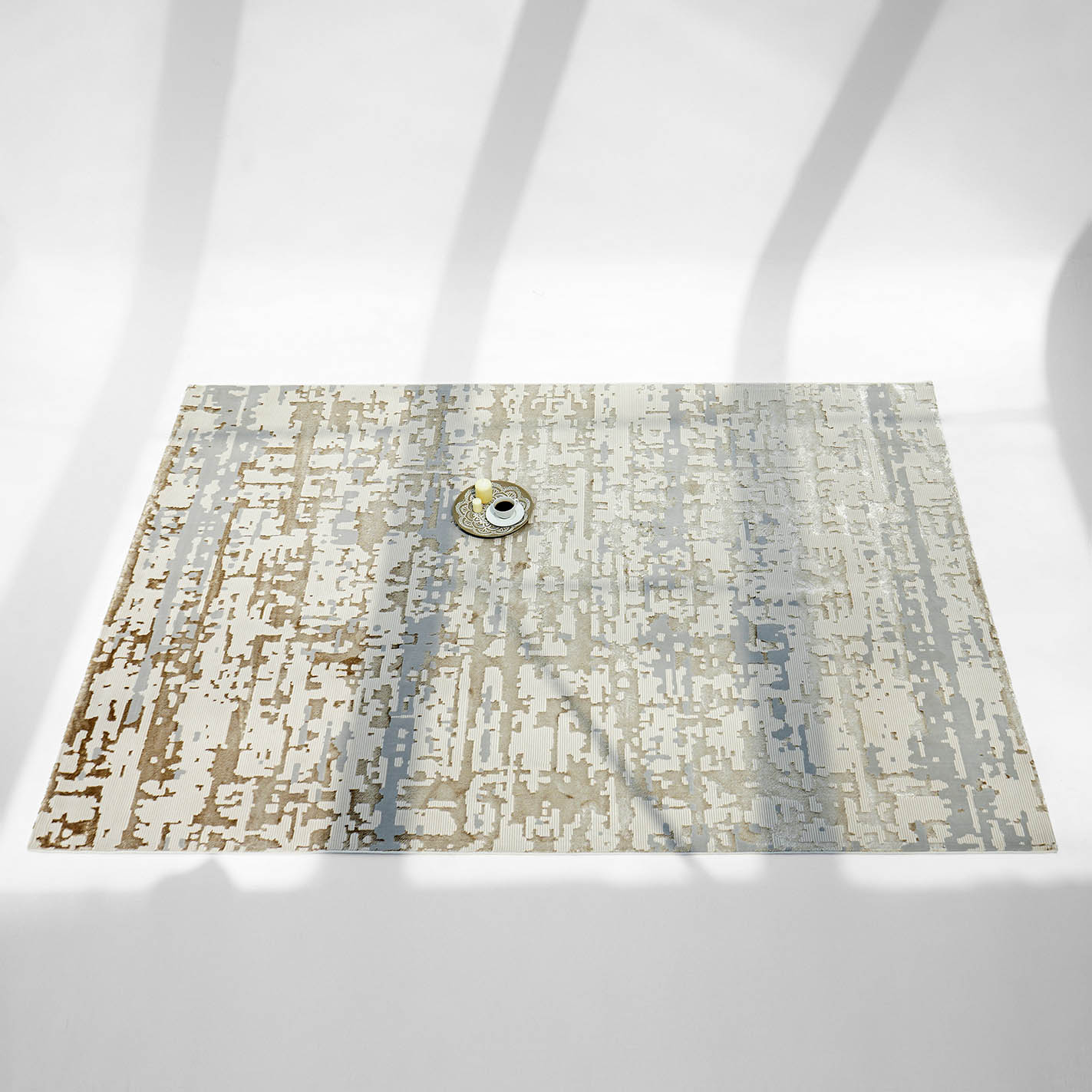 A single cup and saucer rest on the RAMSHA HOME Stunning Abstract Pattern Machine Made Rug - MZ40K, its geometric, neutral-toned design bathed in long shadows cast by objects outside the frame.