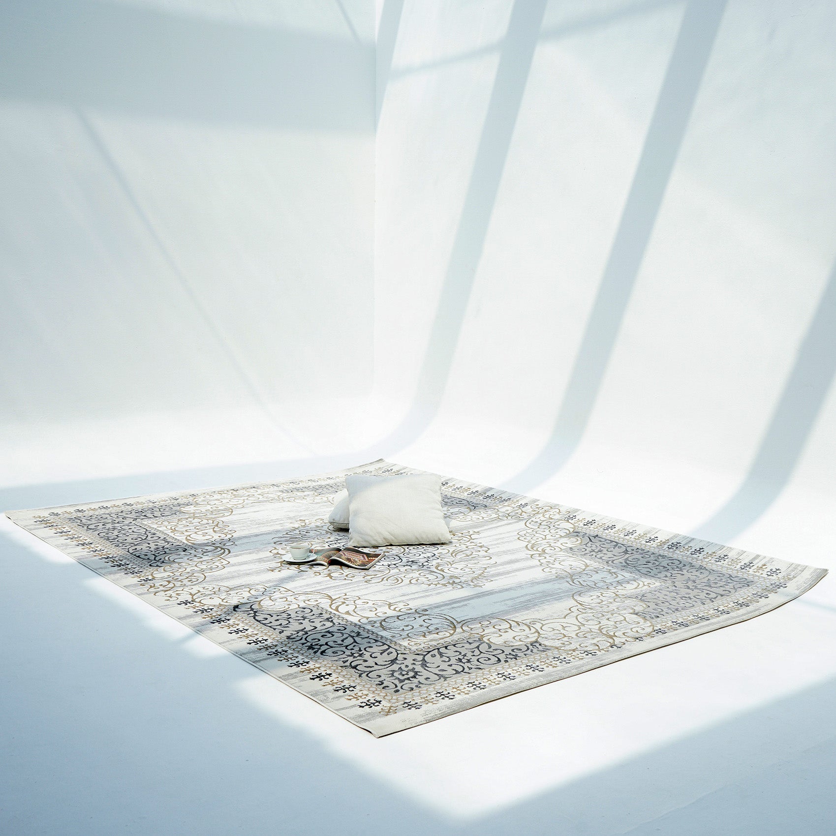 The RAMSHA HOME Light Grey Color NK36B 100% Polyester Silk Rug lies on a white floor in a bright, minimalist room, with a light pillow and eyeglasses resting on it as window shadows fall across the scene.