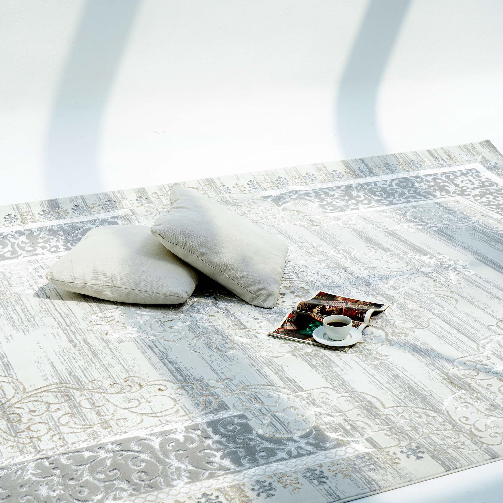 Two light-colored pillows rest on a RAMSHA HOME Light Grey Color NK36B 100% Polyester Silk Rug, beside a coffee cup and saucer, an open book, and sunlight casting soft shadows across the scene.
