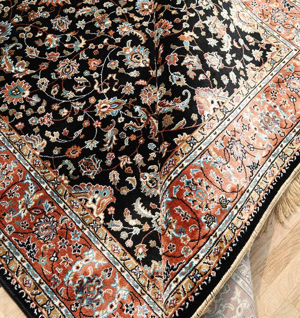 A close-up of the RAMSHA HOME Persian Floral Black Rug with Red Border – MMB 10 (175X275 cm), showing intricate floral patterns on a black background, a red border with motifs, folded corner, and visible fringes and underside.