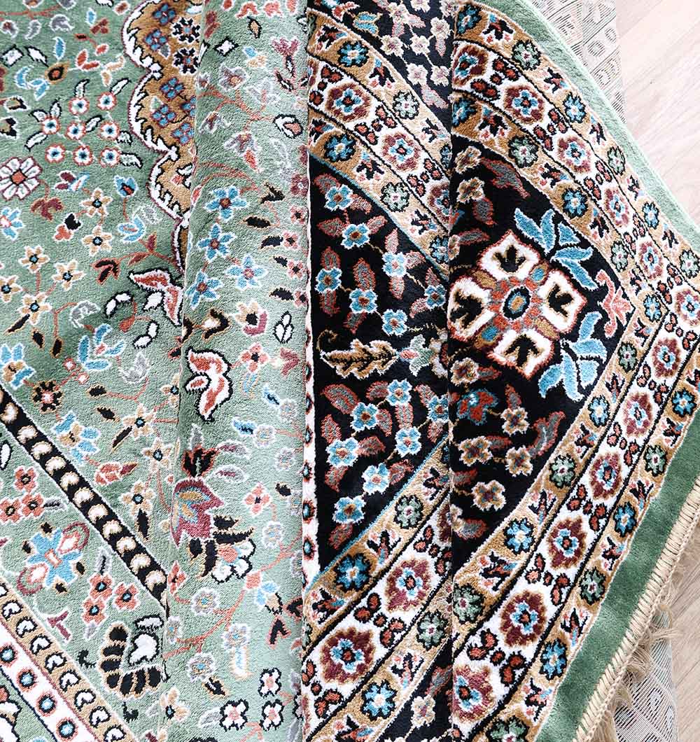 A close-up of the RAMSHA HOME Persian Medallion Mint Green & Black Border Rug – MMB 17 (265x365 cm), showing intricate floral and geometric patterns in green, black, cream, blue, and red, with front and back designs partially visible.