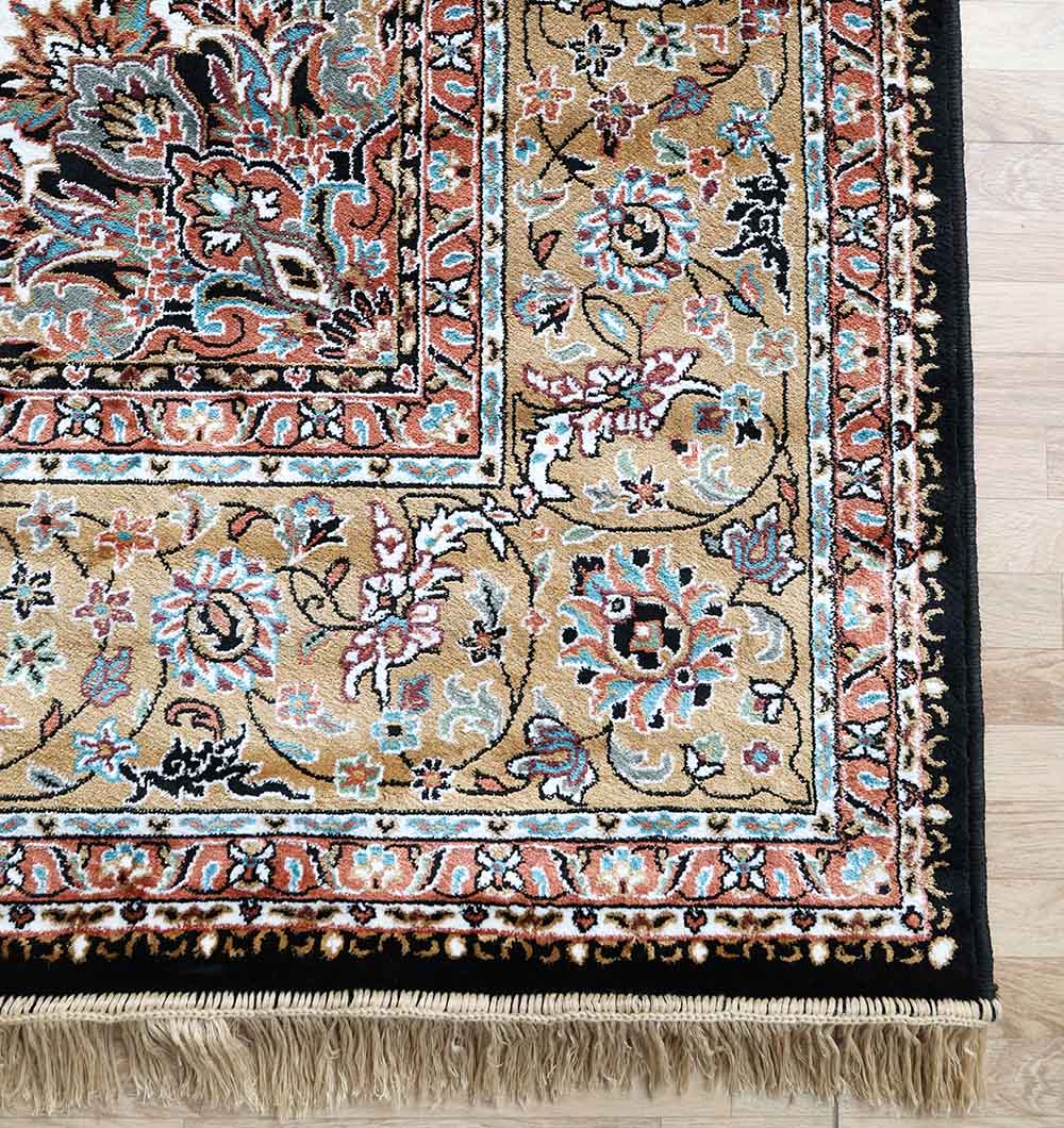 A close-up of the RAMSHA HOME Persian Floral Medallion Black & Gold Rug – MMB 18 (265x365 cm), featuring ornate multicolored floral patterns, a black border, and beige fringe on a light wooden floor.