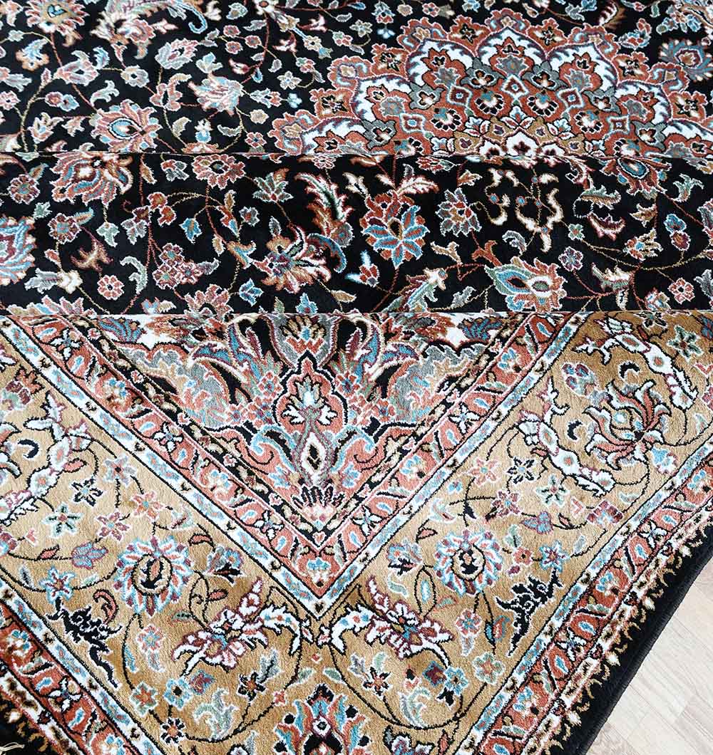 A close-up of the RAMSHA HOME Persian Floral Medallion Black & Gold Rug – MMB 18 (265x365 cm) shows intricate beige, blue, and red floral designs, with one corner folded to reveal its detailed traditional pattern and texture.