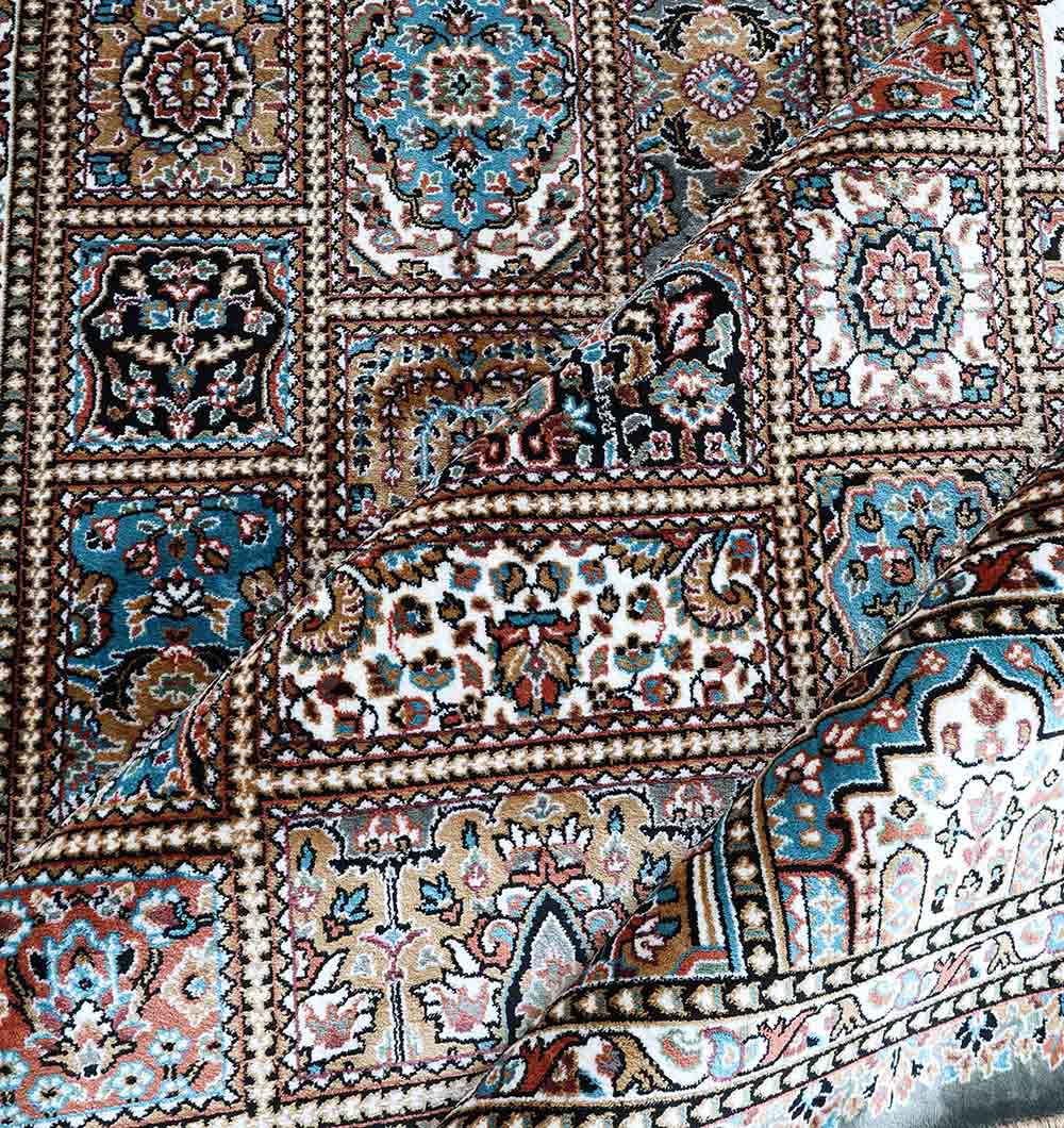 A close-up of the RAMSHA HOME Persian Patchwork Medallion Multicolor Rug – MMB 19 (265X365 cm) showcases intricate blue, brown, beige, and white geometric and floral patterns, with one corner folded to display the underside’s detail.
