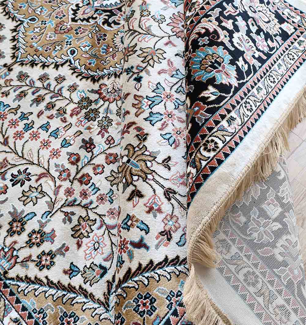 A close-up of the RAMSHA HOME Multi Color Decor Rug - MMB 20 (265X365 cm), showing detailed floral and geometric patterns in beige, black, blue, and pink. One corner is folded to display the fringed edges and the underside of this elegant ivory rug.