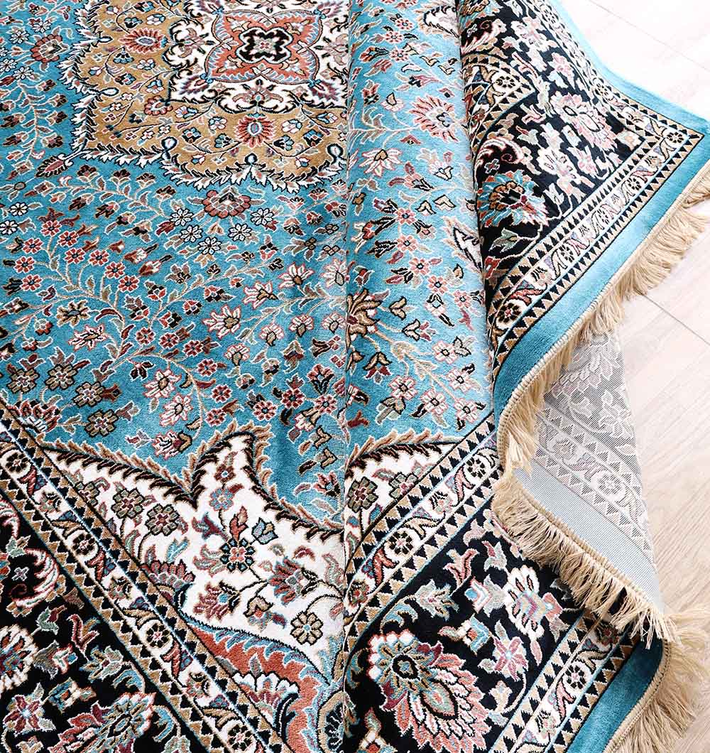 A close-up of the RAMSHA HOME Multi Color Decor Turkish Area Rug - MMB 22 (265X365 cm), showcasing blue, black, cream, gold, and turquoise floral motifs with ornate borders, partially folded to reveal its fringe and underside.
