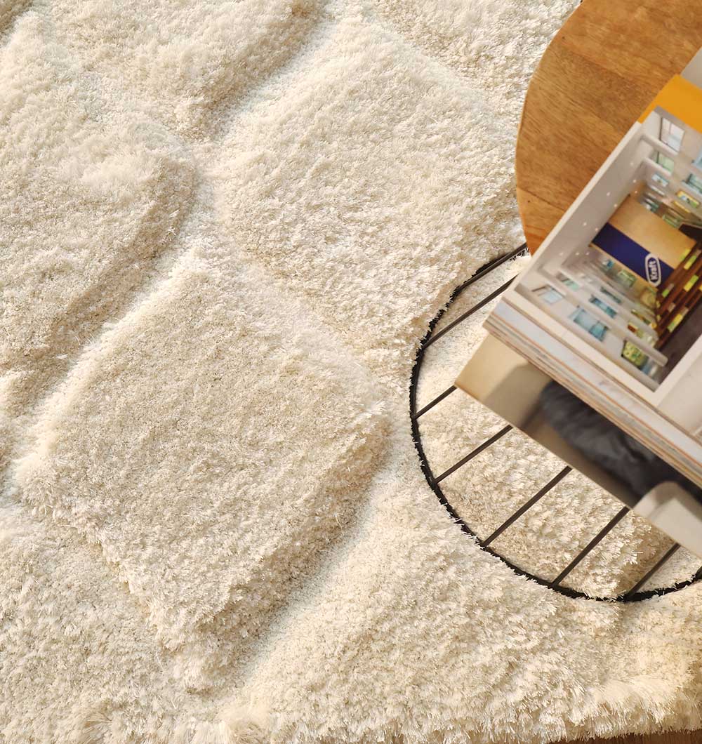 A close-up of the RAMSHA HOME White Rectangle Shaggy Rug with Carved Diamond Pattern (MP3D 13) in ivory, featuring plush pile and raised diamonds, beside a round wooden table topped with stacked magazines.