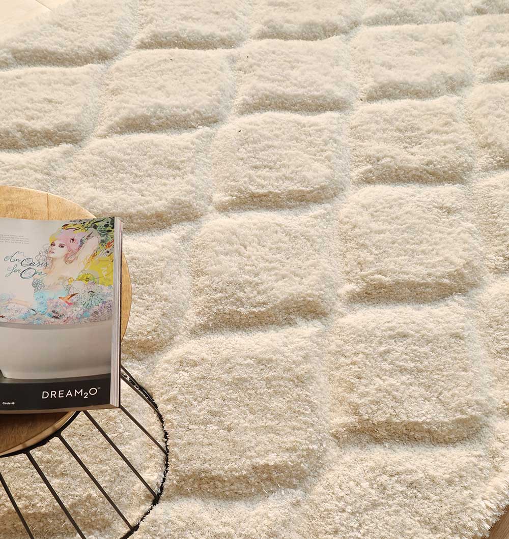 The RAMSHA HOME White Rectangle Shaggy Rug with Carved Diamond Pattern (MP3D 13) features a plush pile and sits beside a round wooden table with an open magazine, while sunlight casts gentle shadows across the scene.