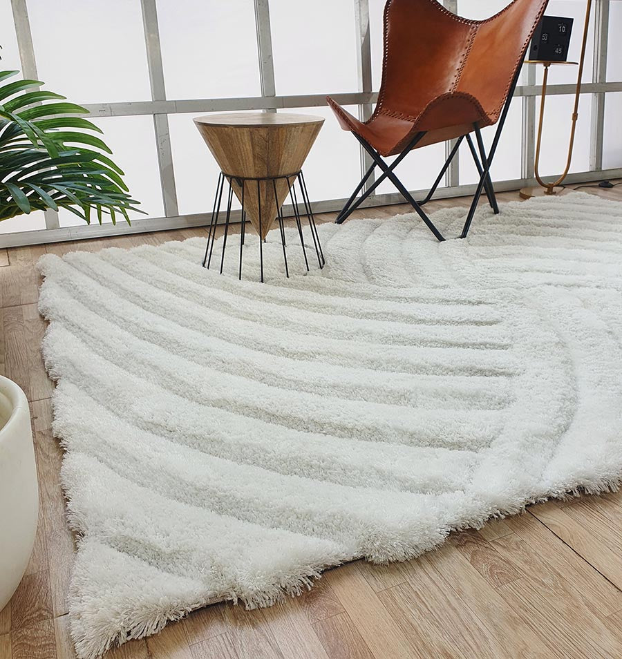 A modern room with a light wood floor, a RAMSHA HOME White Shaggy Area Rug (MP3D 01), a brown leather butterfly chair, a small wooden accent table with black metal legs, and a green plant in a white pot near a large window.