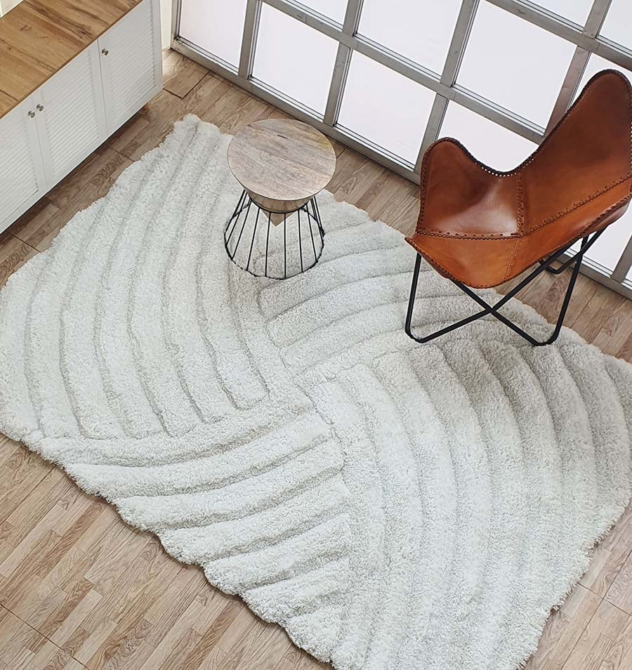 A modern living space with a RAMSHA HOME White Shaggy Area Rug for Large Living Room (MP3D 01), a small round wooden side table with a metal base, and a brown leather butterfly chair on wood floors near large windows.
