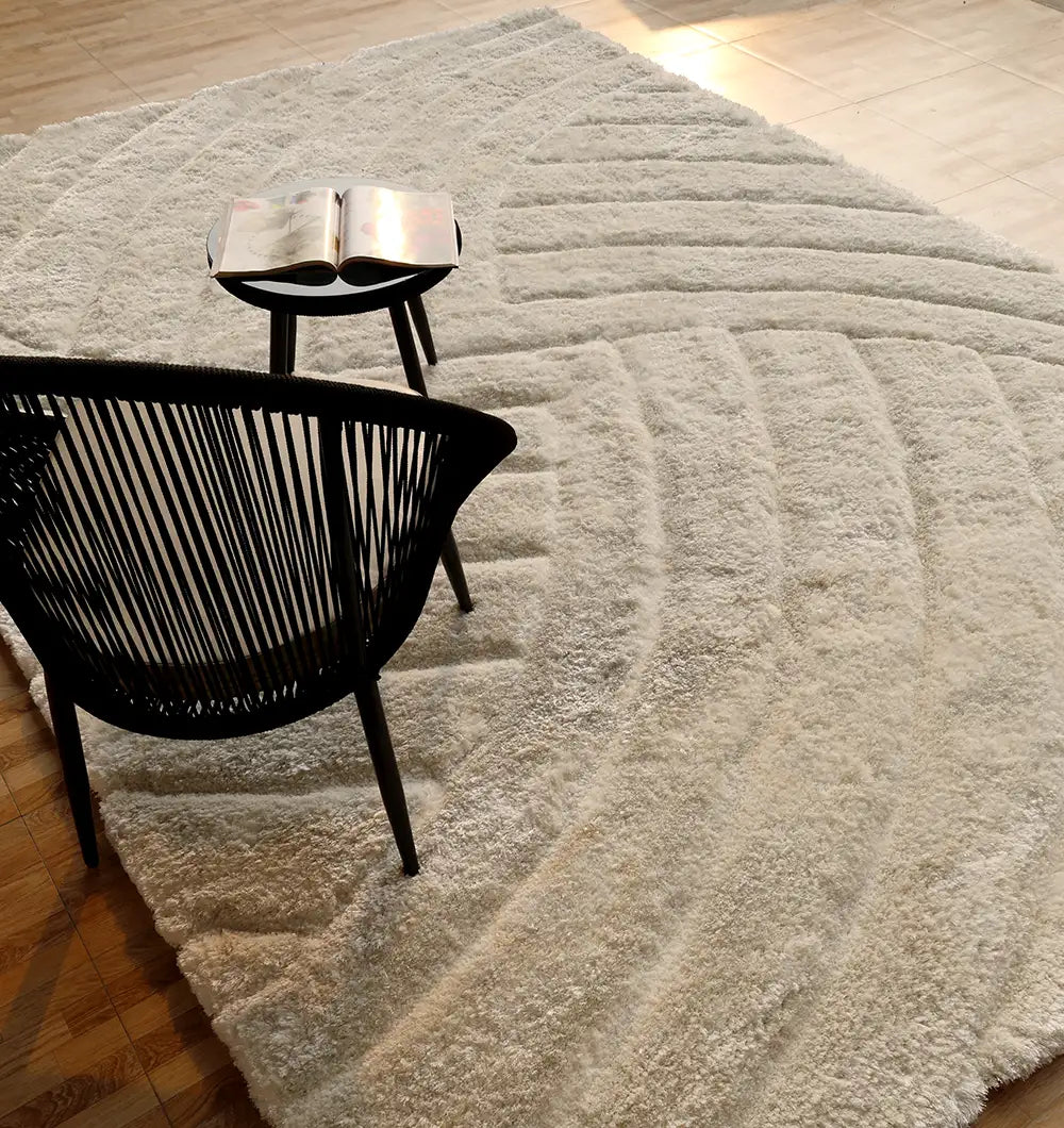 The RAMSHA HOME Textured White Shaggy Area Rug for Large Living Room (MP3D 07) sits on a wooden floor, paired with a black woven chair and round table with an open book, as sunlight partially brightens the cozy scene.