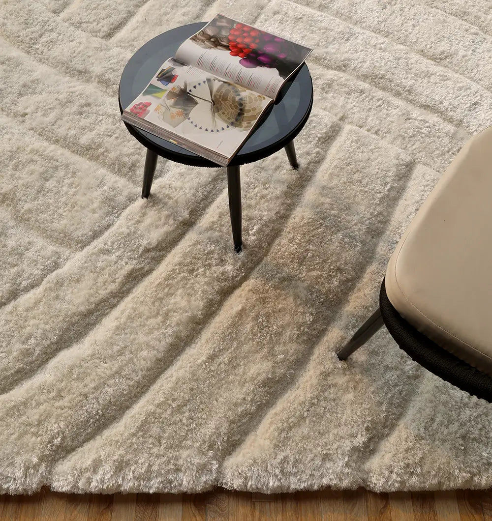 A round black glass coffee table with an open magazine sits on the RAMSHA HOME Textured White Shaggy Area Rug for Large Living Room - MP3D 07, next to a beige cushioned chair on a wooden floor.