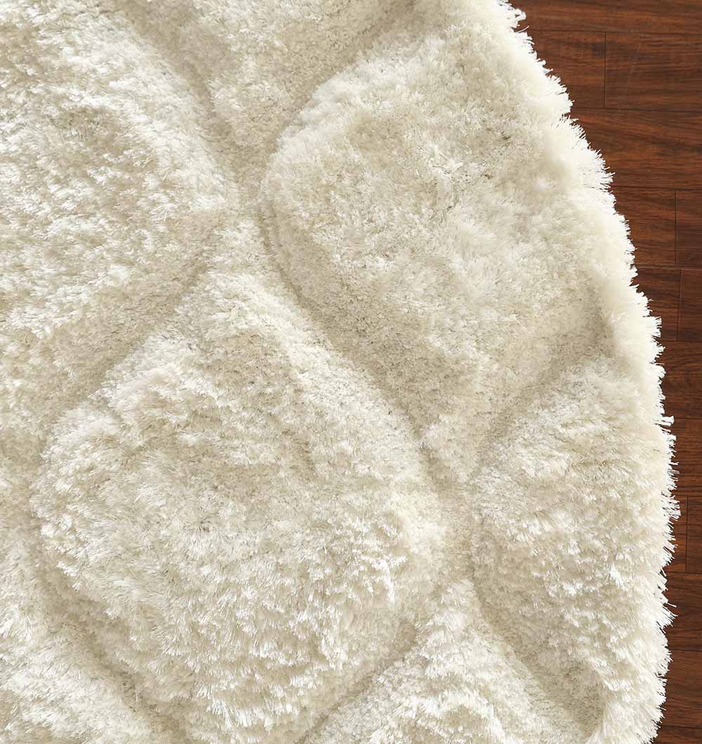 A close-up of the RAMSHA HOME White Round Shaggy Rug with Carved Pattern (MP3D 13) highlights its plush, ivory texture and subtle design, adding soft comfort and style to a dark wooden floor.