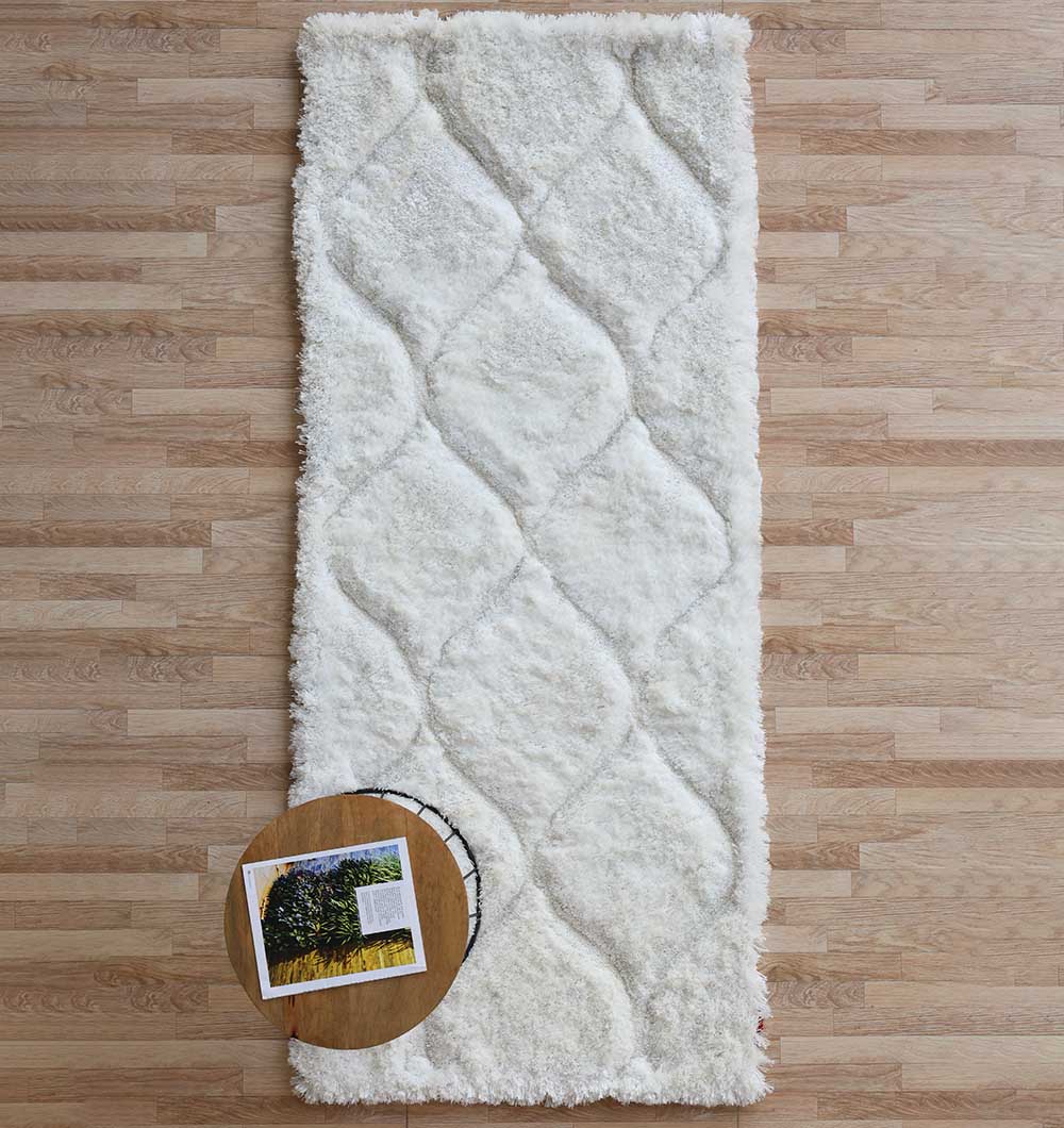 A RAMSHA HOME White Rectangle Shaggy Rug with Carved Diamond Pattern (MP3D 13) lies on a light wooden floor, while a round side table in the corner holds an open book and a photograph.