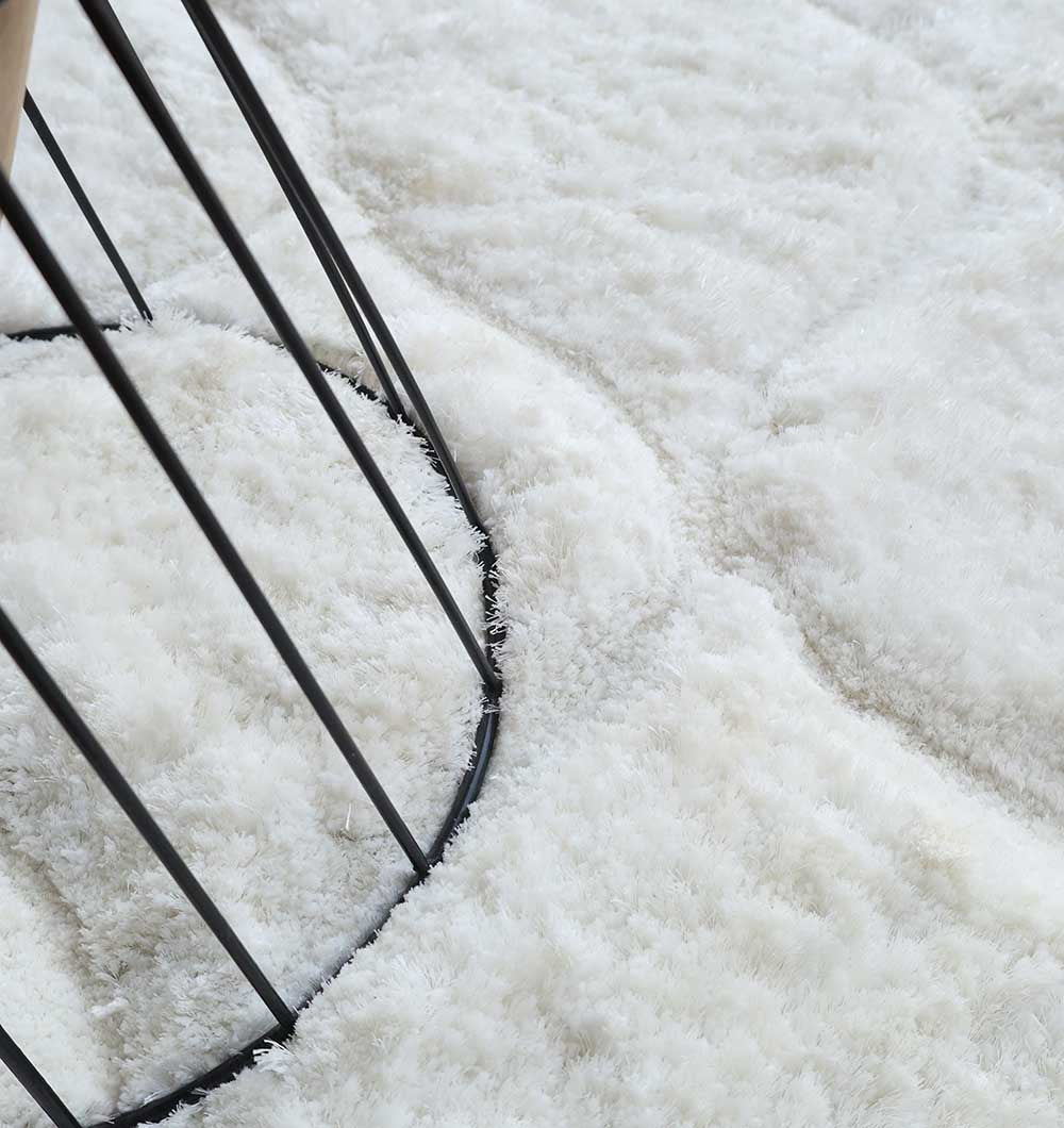 Close-up of the RAMSHA HOME White Rectangle Shaggy Rug with Carved Diamond Pattern - MP3D 13, showcasing its plush pile beside a round black metal table base and highlighting the contrast between the soft rug surface and hard metal bars.