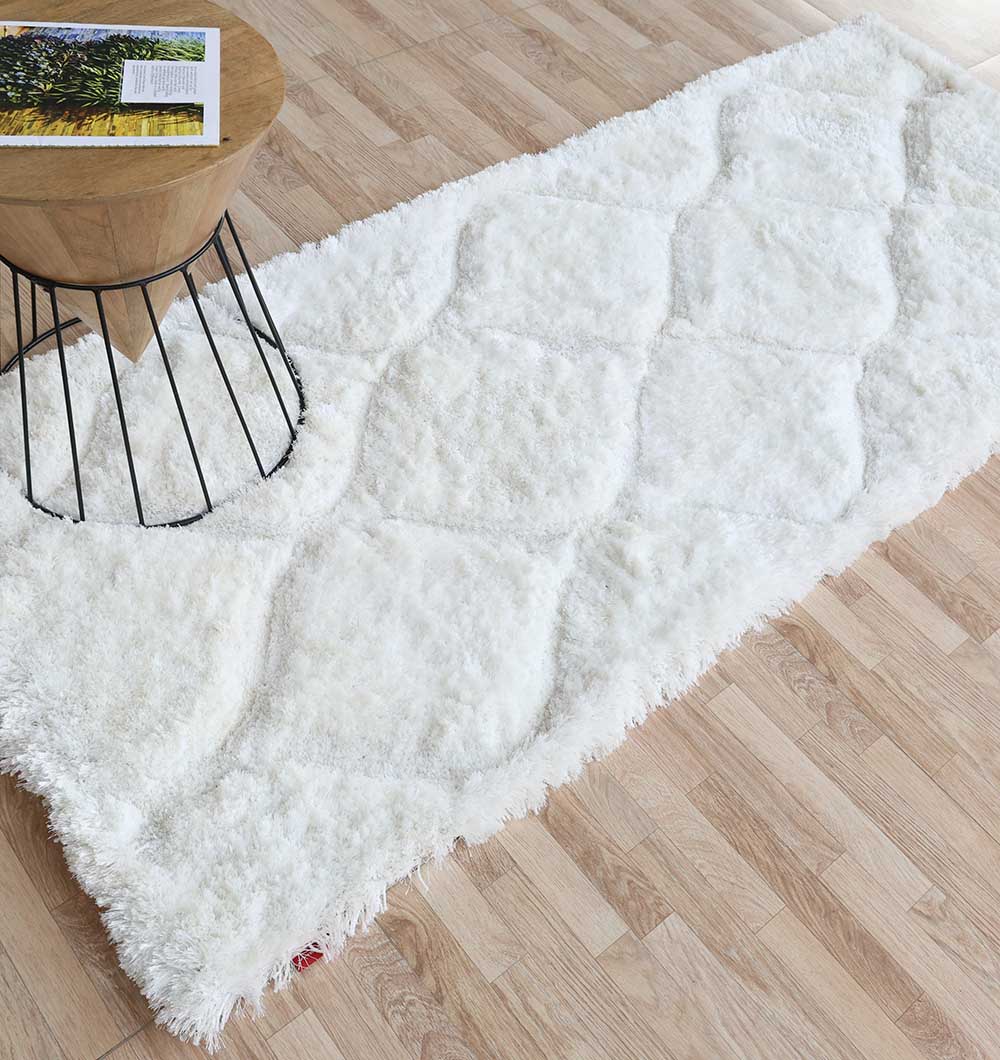 The RAMSHA HOME White Rectangle Shaggy Rug with Carved Diamond Pattern (MP3D 13) lies on a light wood floor beside a small round wooden table with a black wire base, topped with a book and magazine.