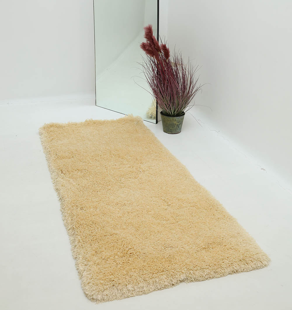 The Handmade Fluffy Shaggy Rug, Cream Color - MPV 08 is placed on a white floor beside a potted plant with long burgundy leaves and a large mirror, all against a plain white wall for a cozy, solid color rug look.