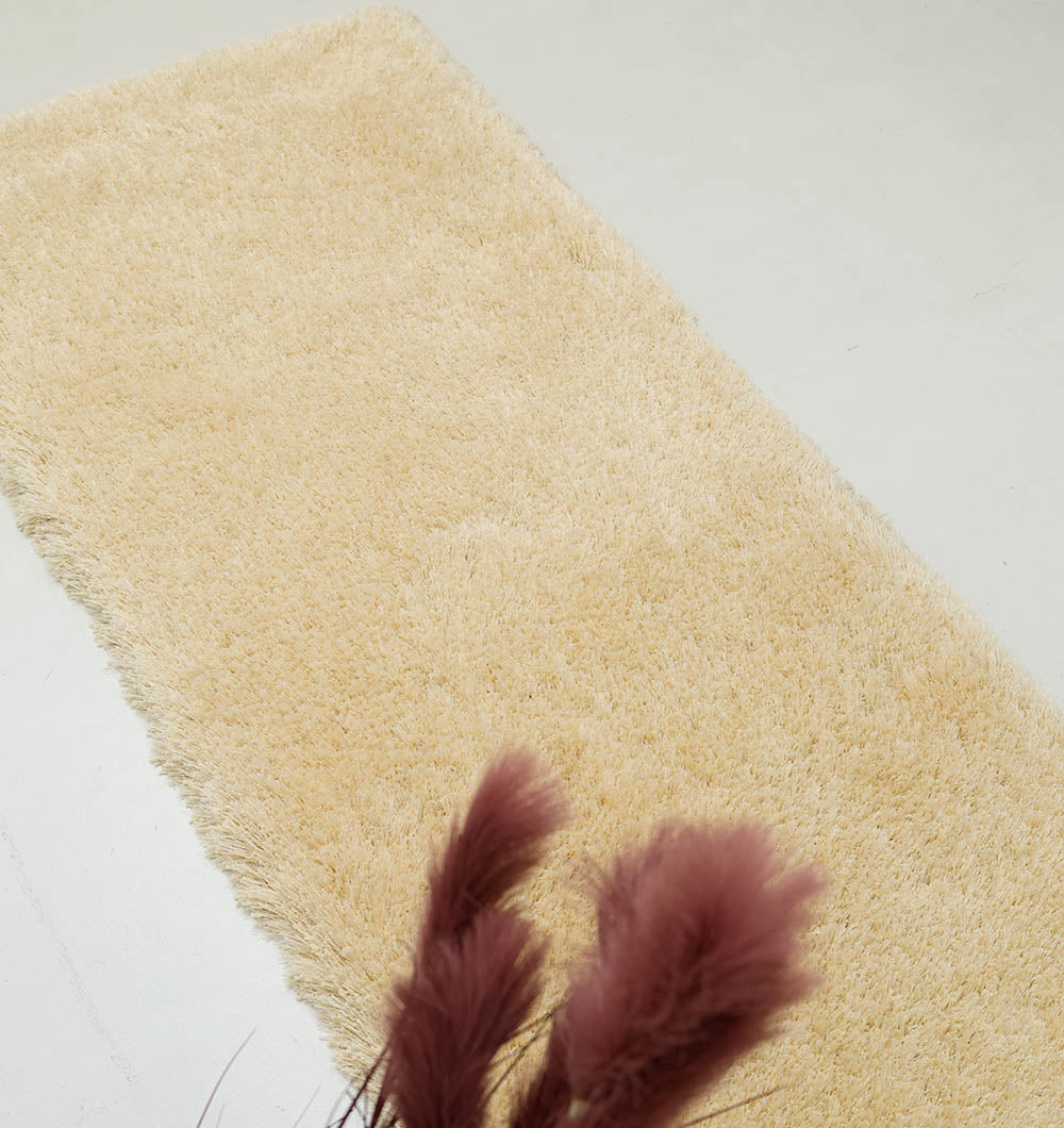 A Handmade Fluffy Shaggy Rug in cream (MPV 08) lies on a light floor. Out of focus in the foreground, burgundy dried grass adds contrast to the rug’s soft, cozy texture and solid color.