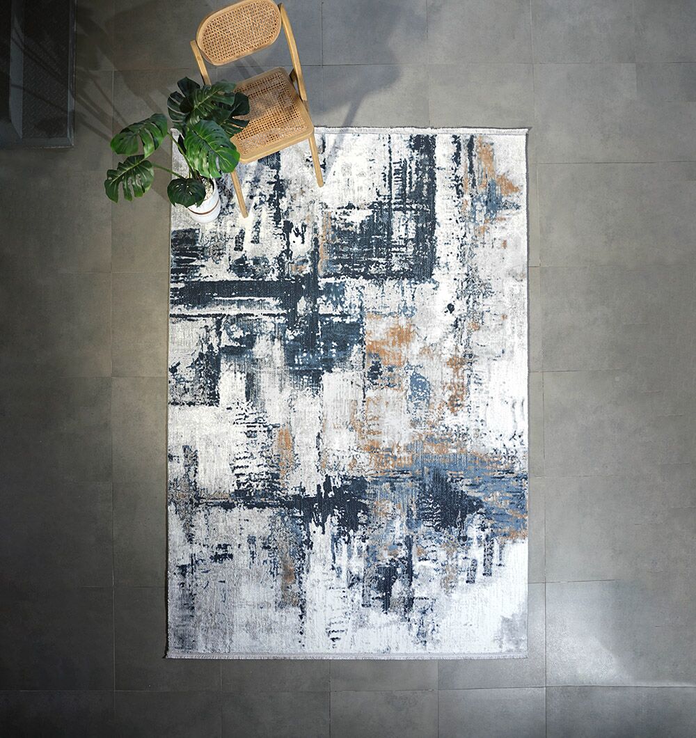 The RAMSHA HOME Machine Made Turkish Rug - MR112 FLORENCE, featuring abstract blue, gray, and beige patterns, is spread on a gray tiled floor with a wooden chair and potted plant at one corner.