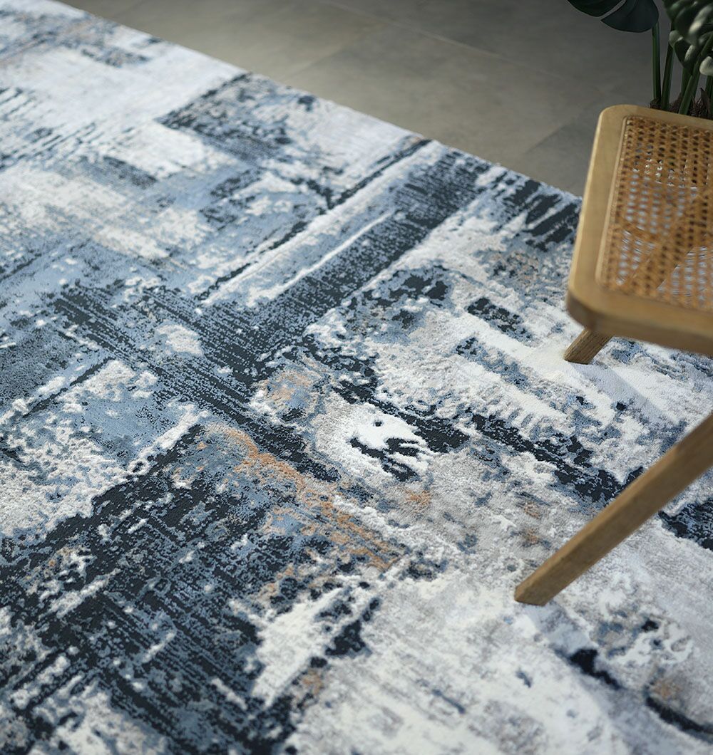 A close-up of the RAMSHA HOME Machine Made Turkish Rug - MR112 FLORENCE, featuring an abstract blue, gray, and white pattern. A wooden chair with a woven rattan seat appears in the upper right on the tiled floor.