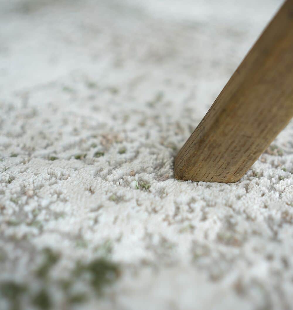 Close-up of a wooden furniture leg pressing into the Cream Green Machine Made Turkish Rug - MR165 FLORENCE by RAMSHA HOME, featuring soft, textured fibers in light cream and soothing sage green tones.