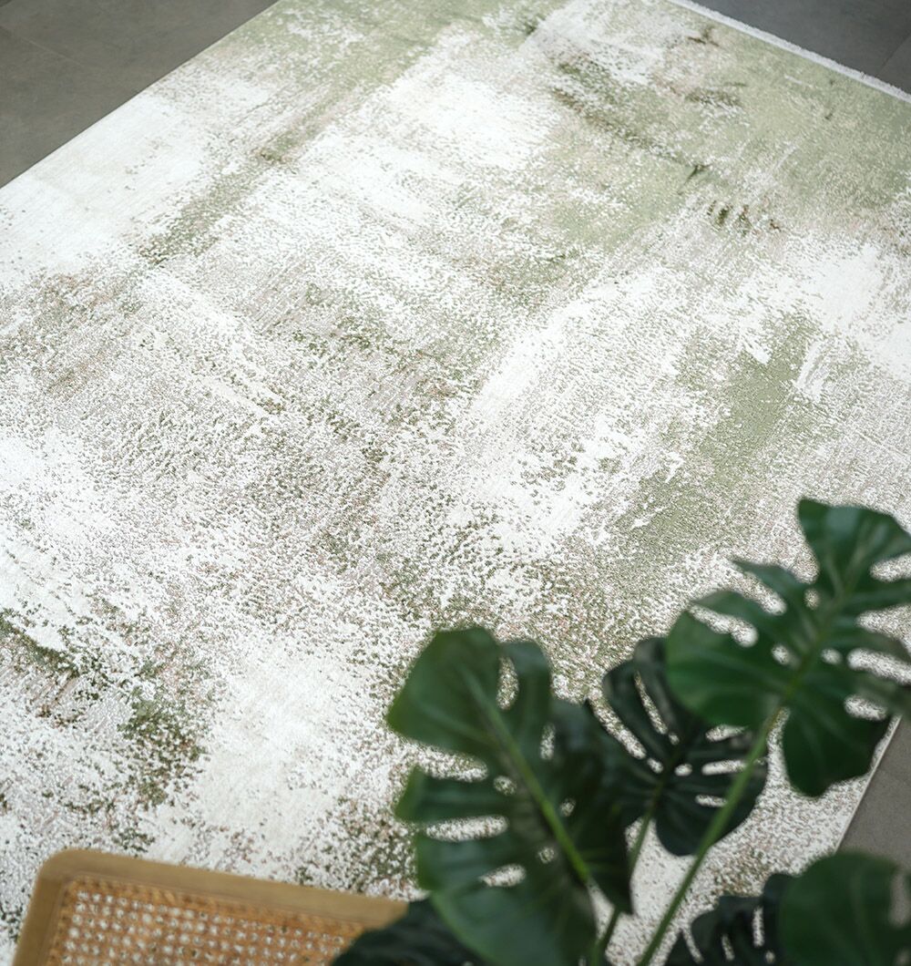 The RAMSHA HOME Cream Green Machine Made Turkish Rug - MR165 FLORENCE, featuring a subtle abstract green and white pattern, is placed on the floor with a green leafy plant and part of a woven chair visible in the foreground.