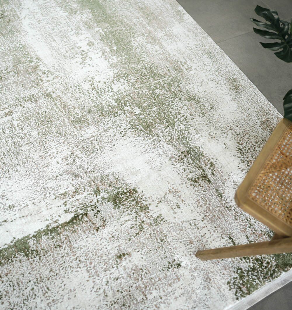 The RAMSHA HOME Cream Green Machine Made Turkish Rug - MR165 FLORENCE, featuring a mottled pattern in white, beige, and sage green, rests on a light tile floor near a wooden chair with a woven seat and a vibrant green plant.