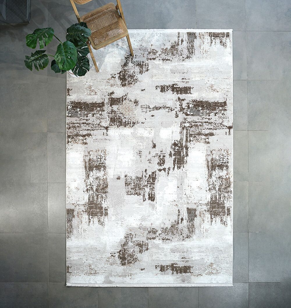 The RAMSHA HOME MR167 FLORENCE Turkish area rug, featuring a modern distressed ivory design with brown and white abstract patterns, is placed on gray tiles with a wooden chair and leafy green plant at its top.