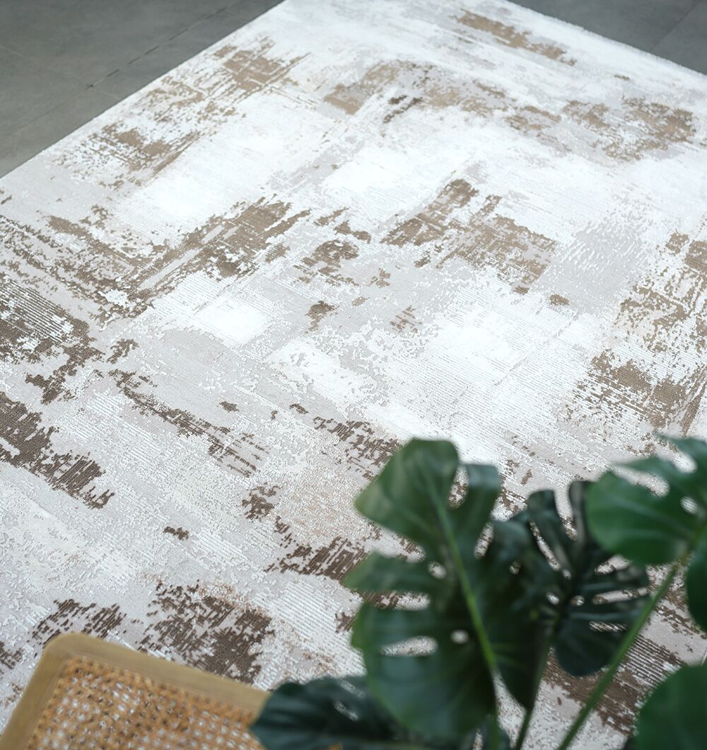 The RAMSHA HOME MR167 FLORENCE Turkish area rug, featuring a modern abstract beige and white pattern, is laid on the floor with a green leafy plant and the corner of a woven chair in the foreground.