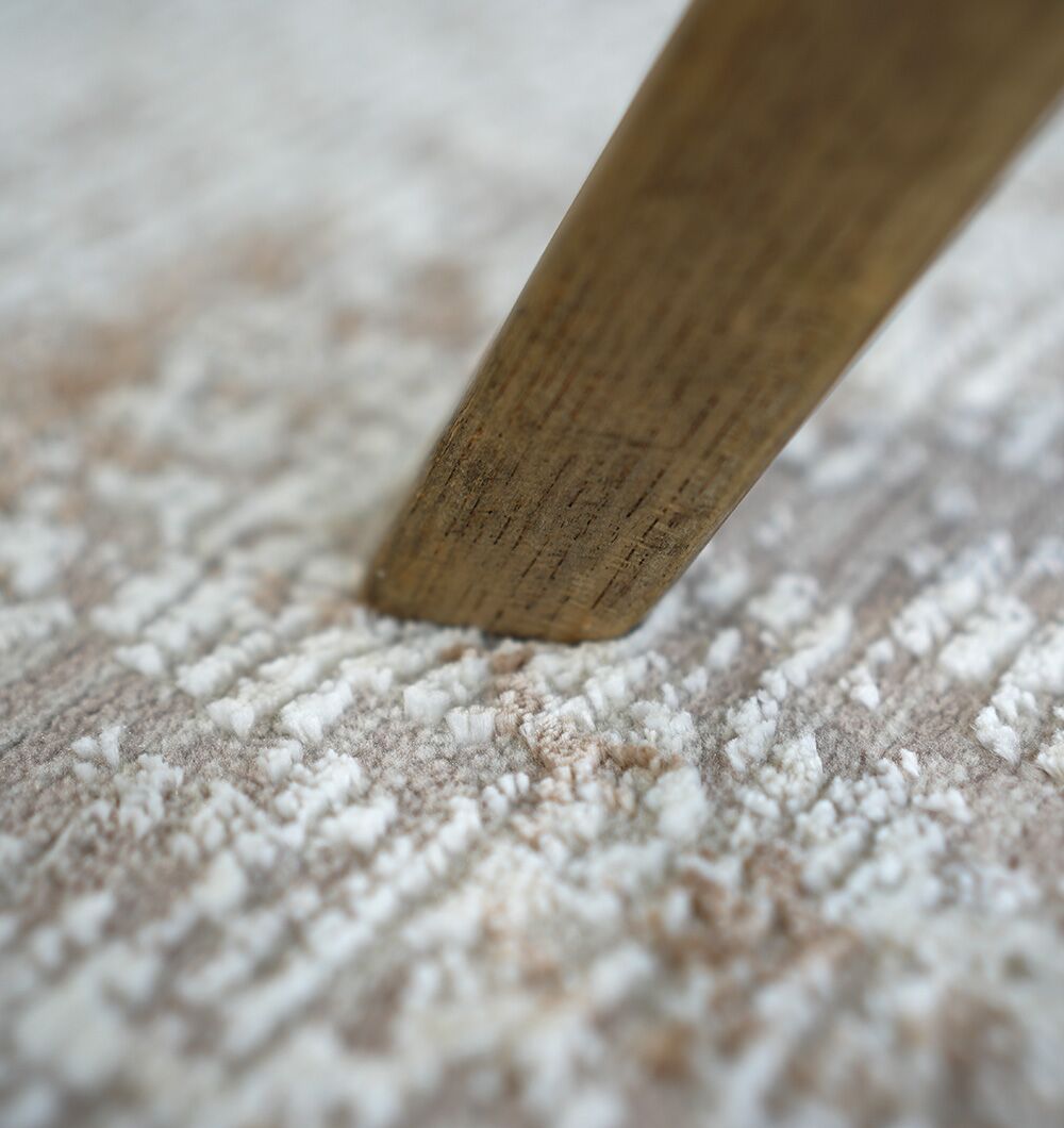 A close-up of a wooden furniture leg resting on the RAMSHA HOME Cream Gold Machine Made Turkish Rug (MR172 FLORENCE), featuring a light ivory color and subtle abstract pattern.